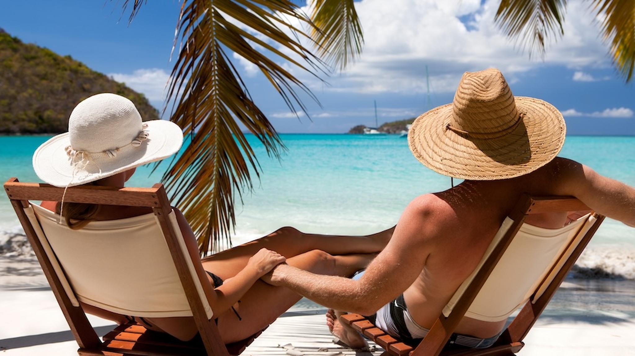 A couple relaxes on lounge chairs under palm trees, gazing at a turquoise sea and clear sky on a sunny beach.