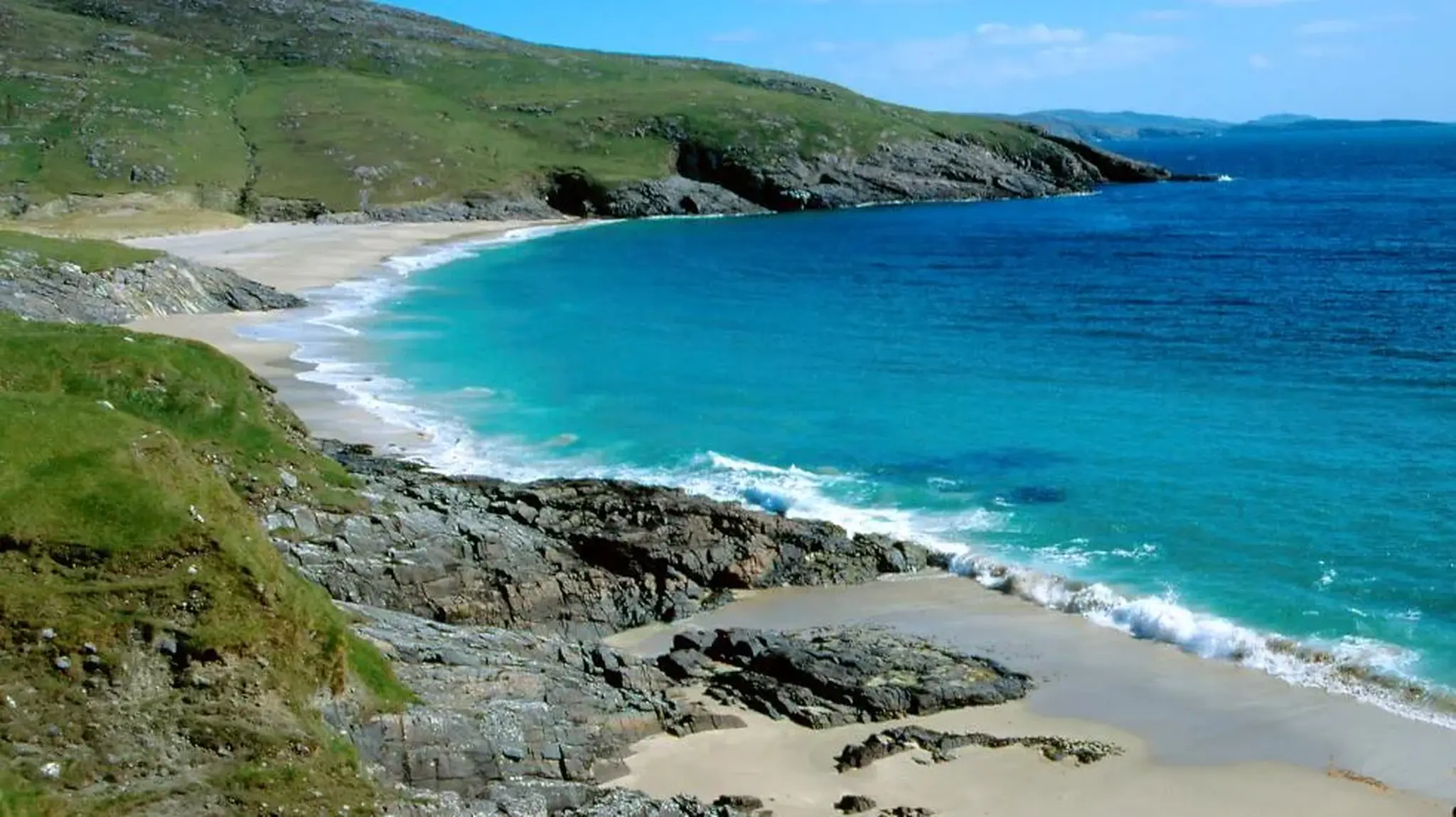 A scenic coastal view with turquoise water, sandy beaches, rocky cliffs, and grassy hills
