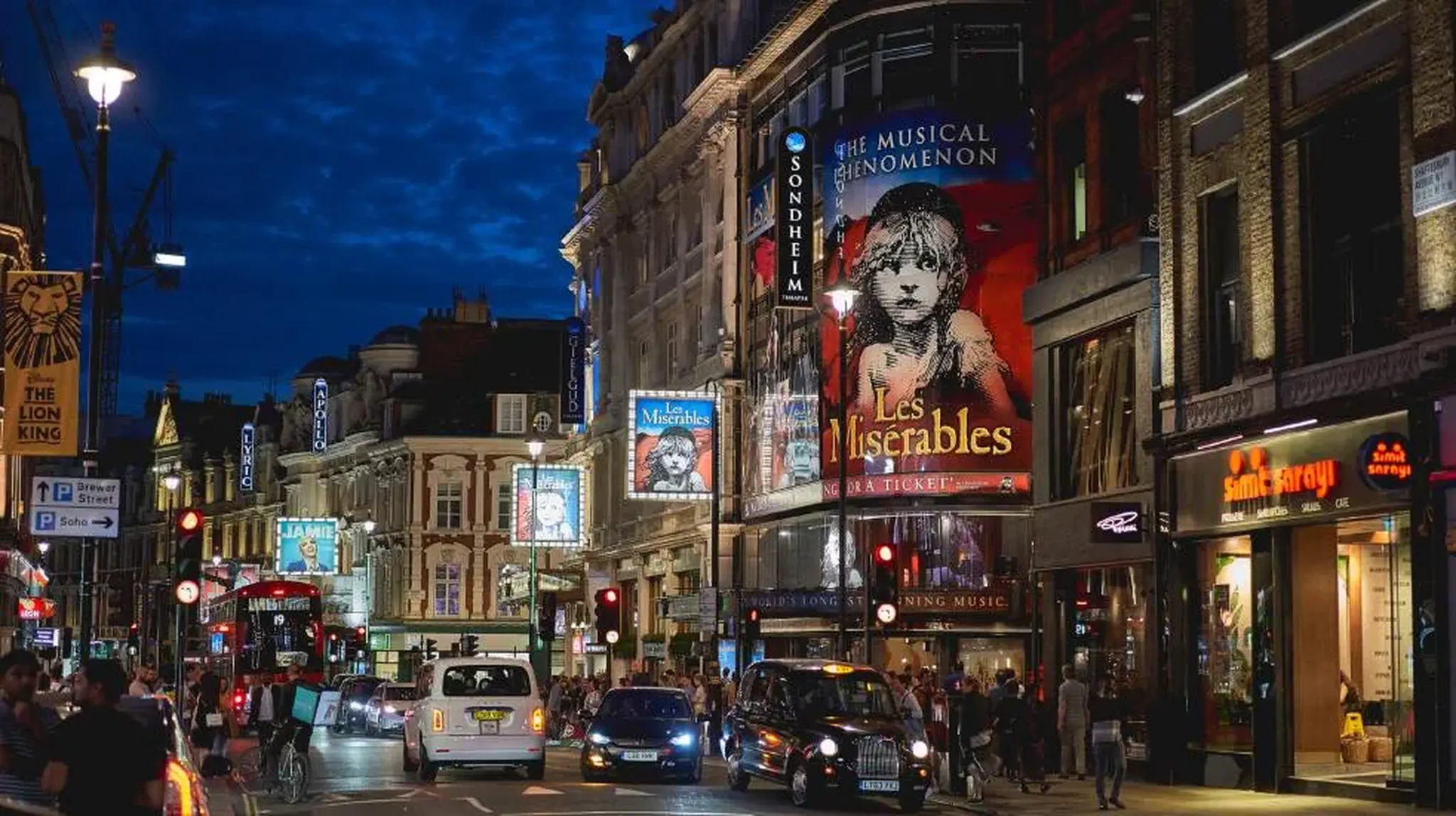 Busy London street at night, illuminated with theatre signs saying "Les Misérables."