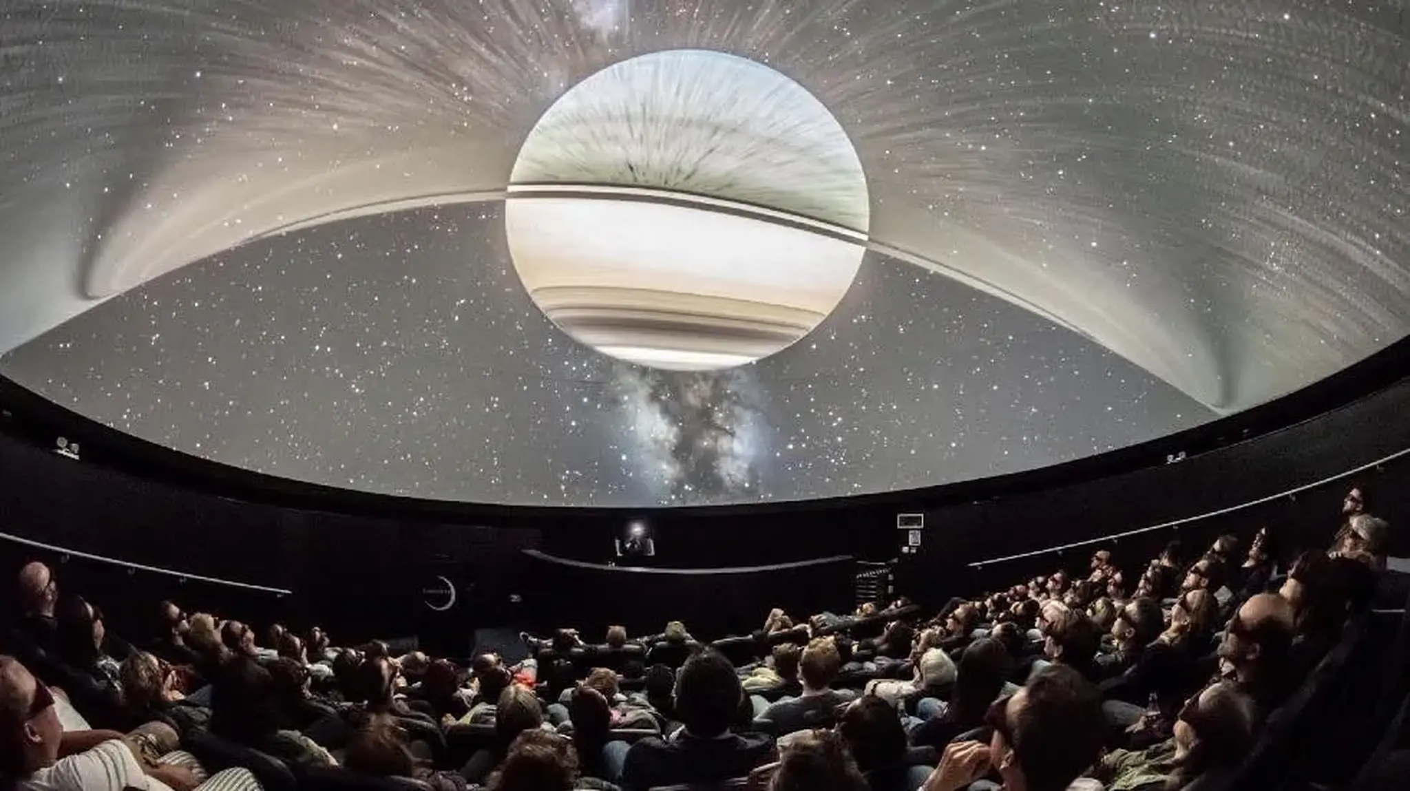 People seated in a planetarium, gazing at a large, detailed projection of Saturn