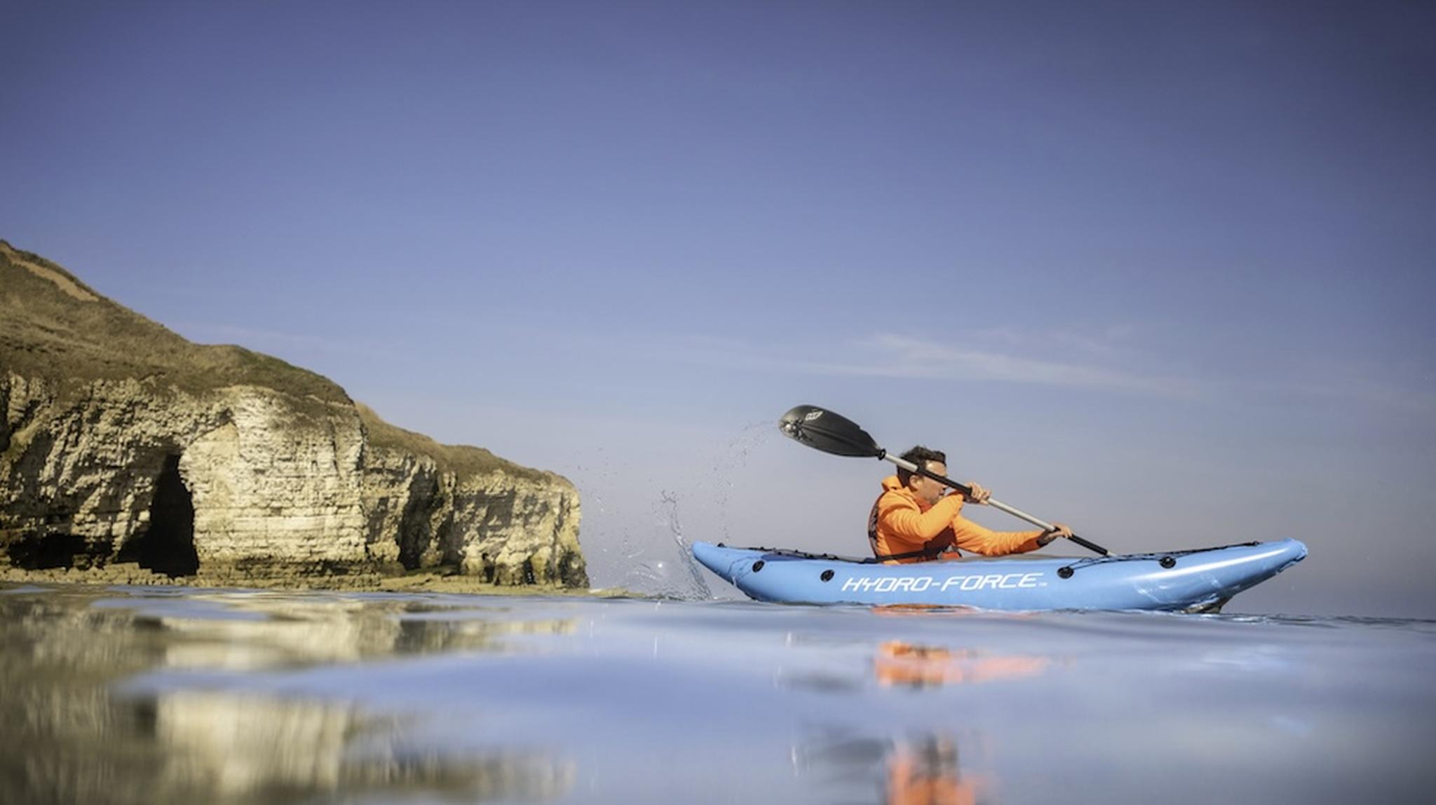 Person in an orange jacket kayaking on calm sea near rocky cliffs under a clear blue sky.
