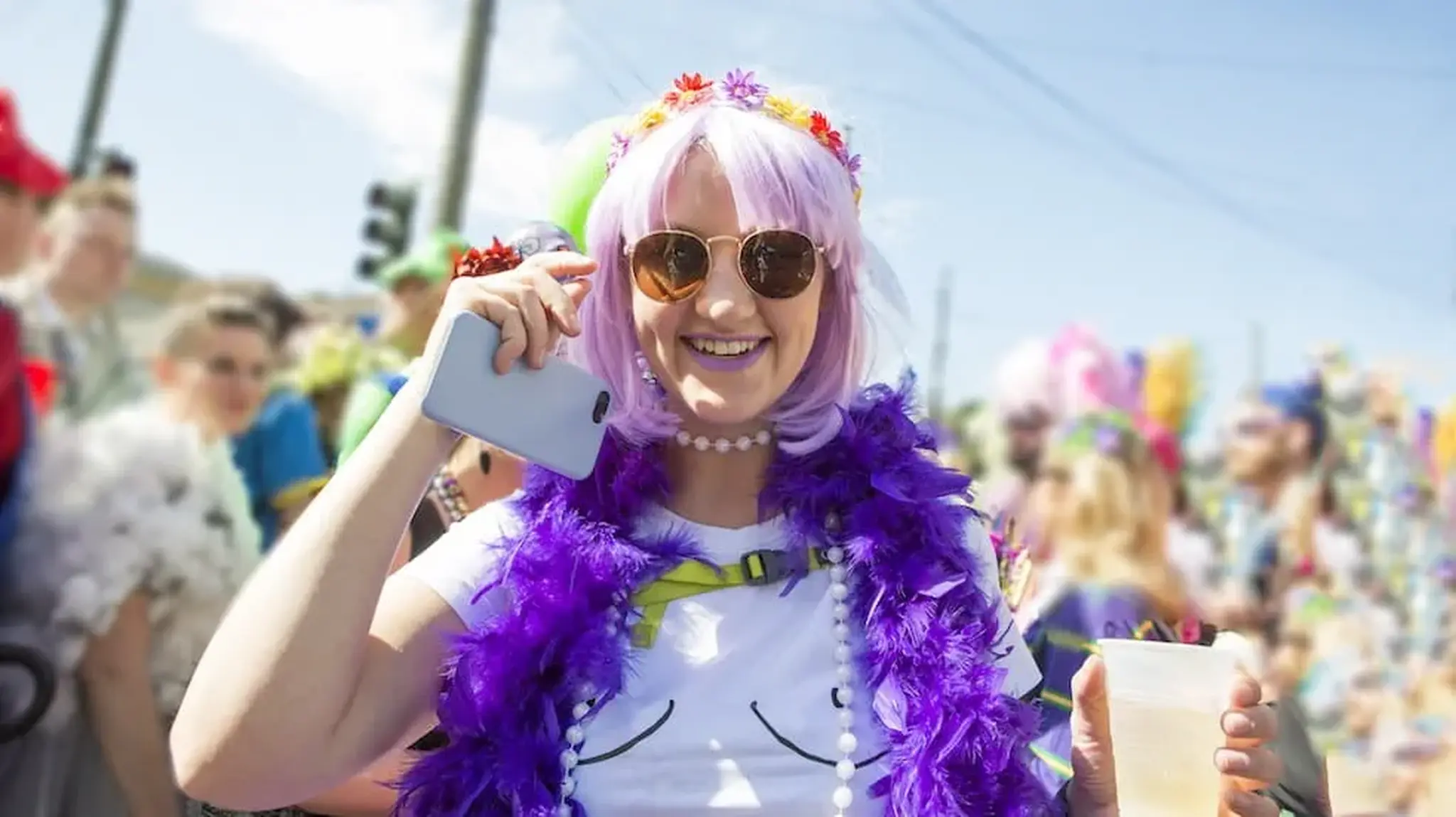 Person in purple wig and sunglasses smiling, wearing a floral headband and feather boa