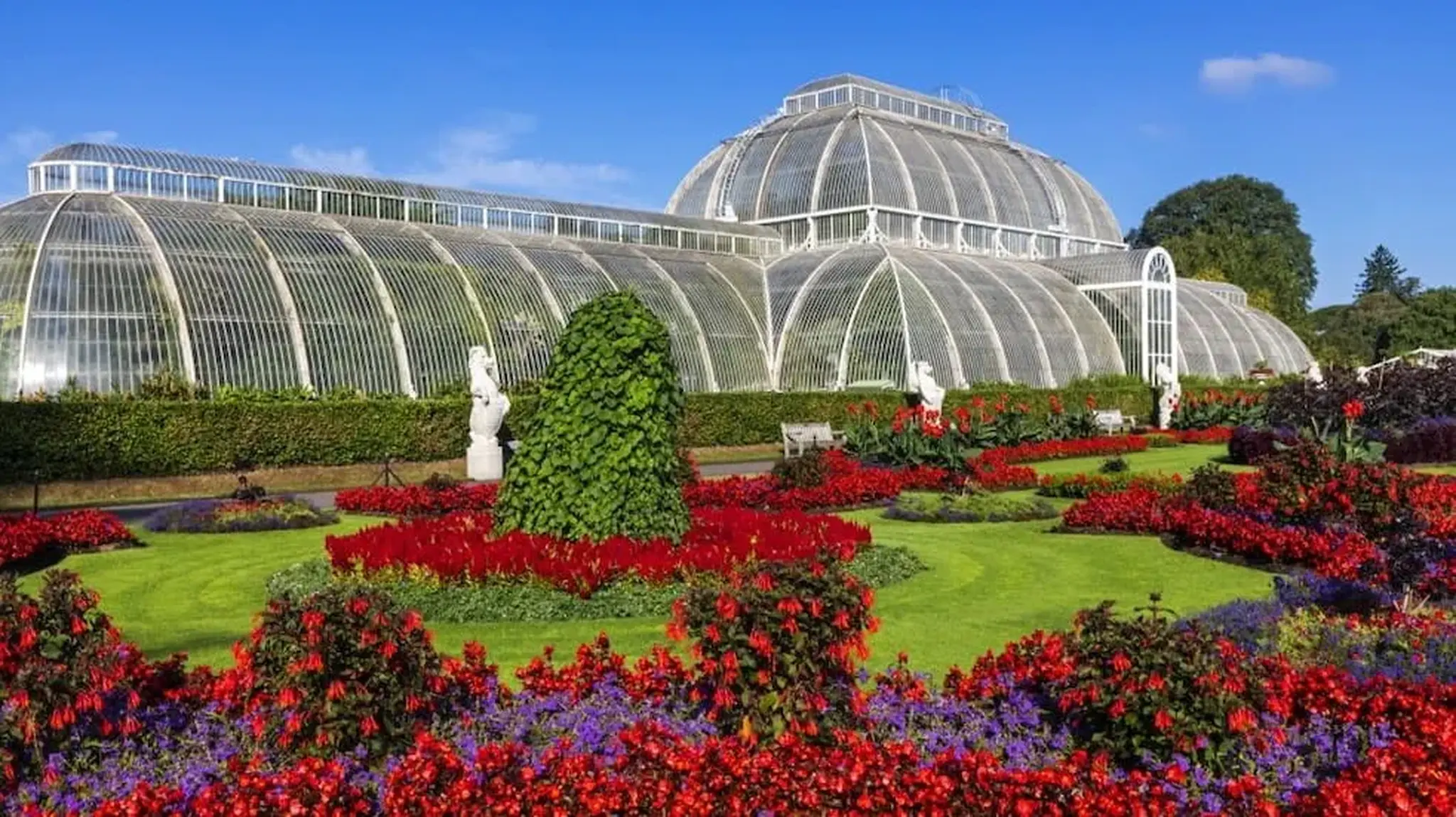 A large glass greenhouse with ornate architecture is surrounded by vibrant red and purple flower gardens