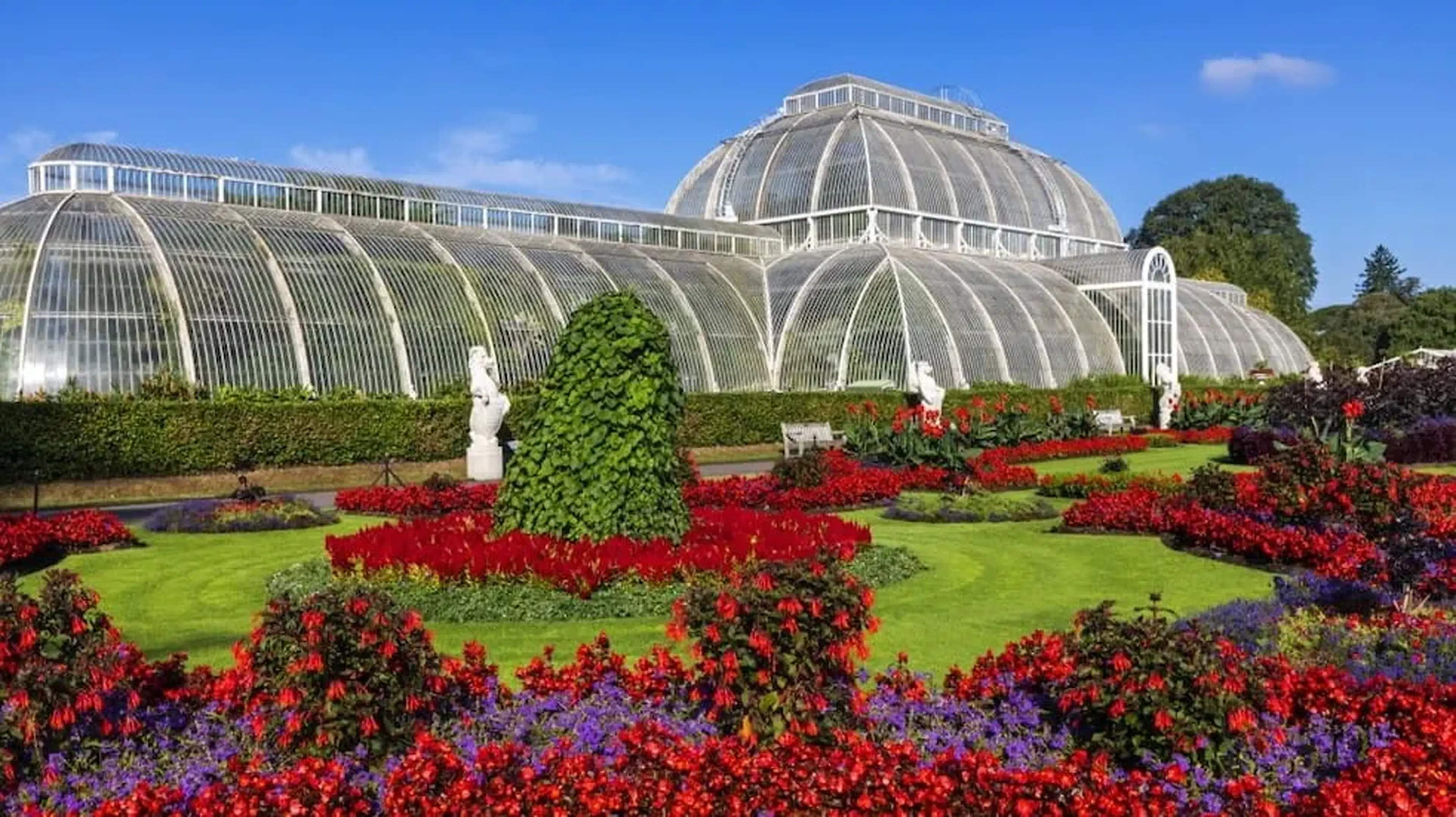 A large glass greenhouse with ornate architecture is surrounded by vibrant red and purple flower gardens