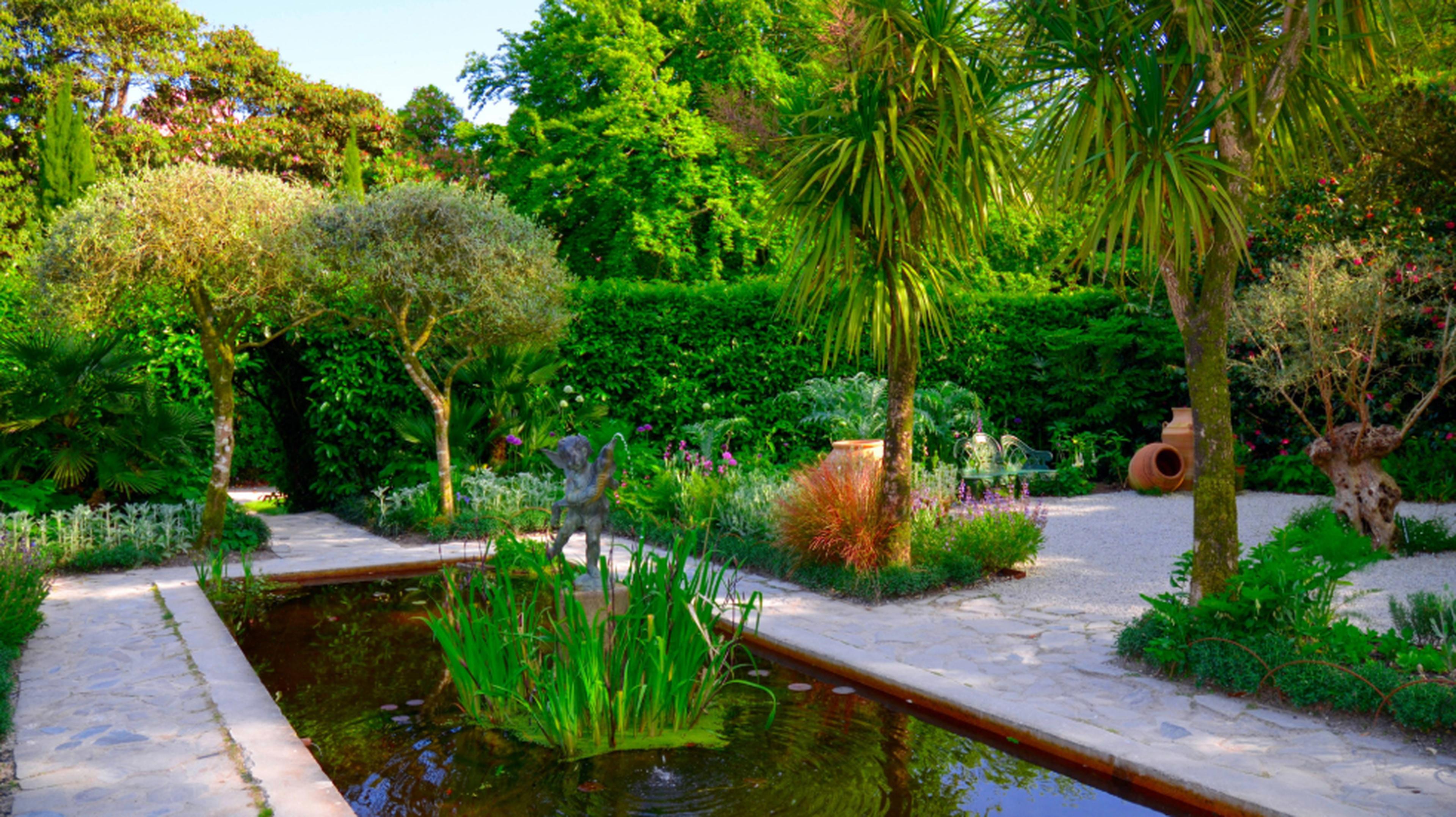 Lush garden with a stone pathway, palm trees, a small rectangular pond with greenery, and a statue in a serene setting.