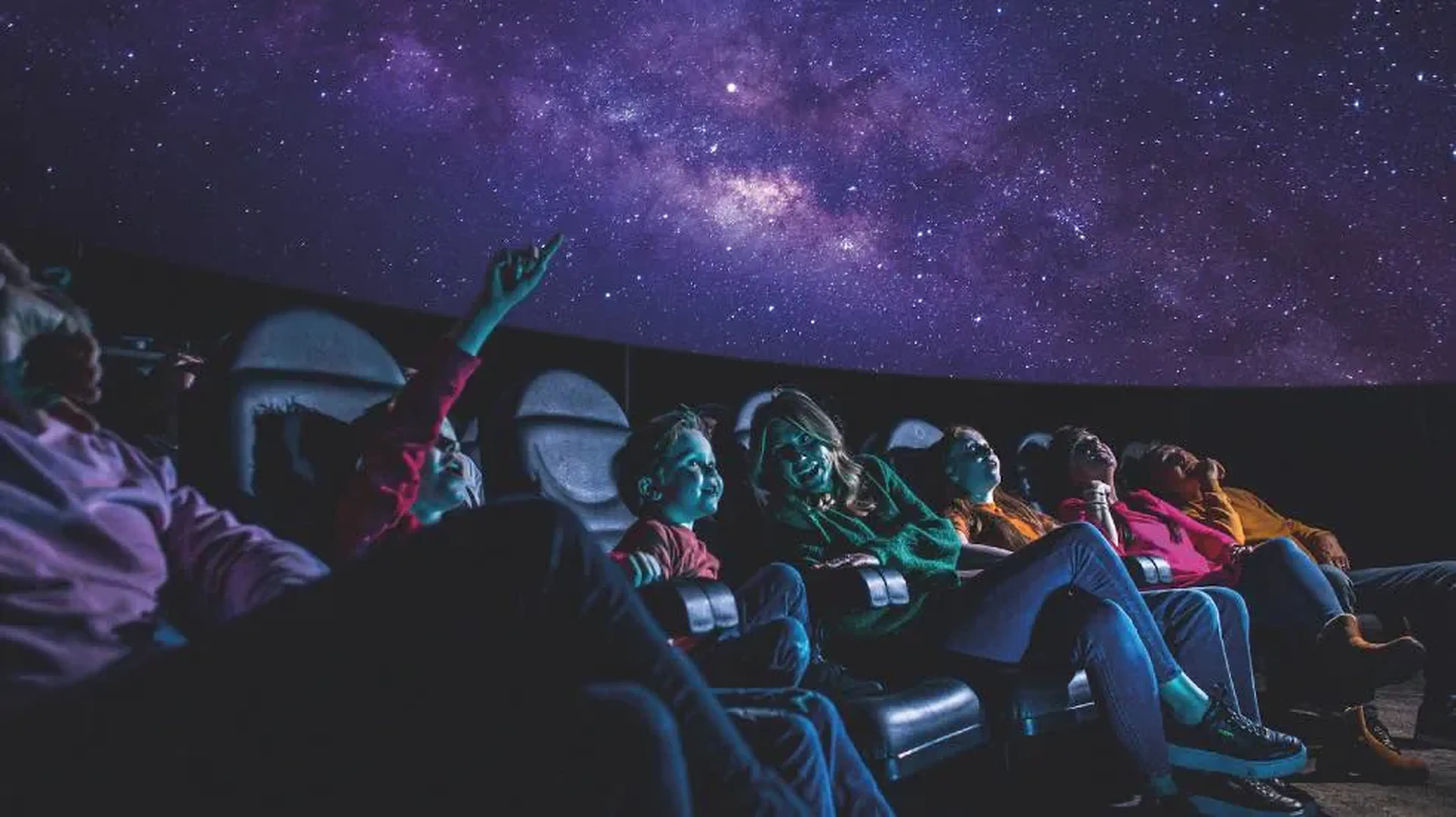 People in reclined seats gaze at a dome-shaped planetarium ceiling displaying a star-filled night sky.