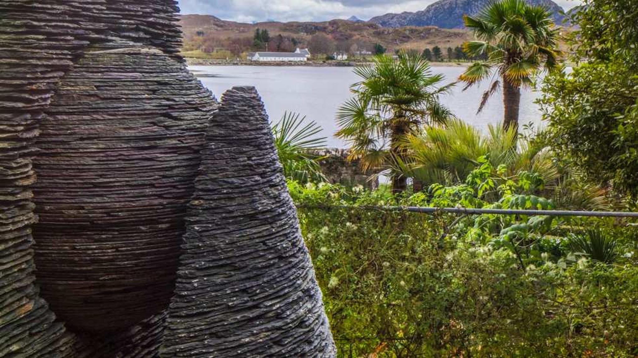 Stone sculpture with spiral texture in foreground, greenery, palm trees, and lake