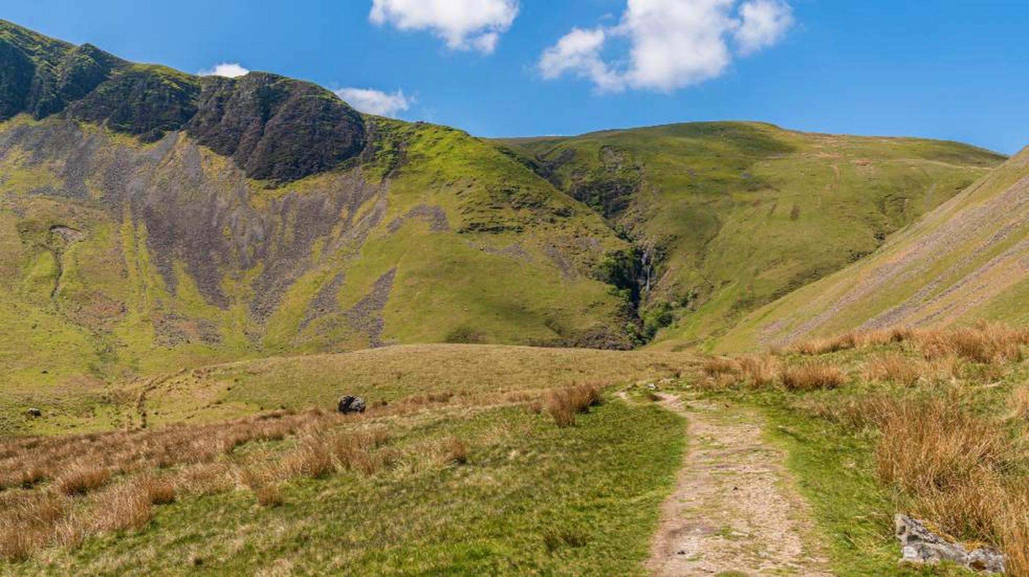Mountain landscape with grassy hills, a pathway leading through the valley, and bright blue sky with a few clouds.