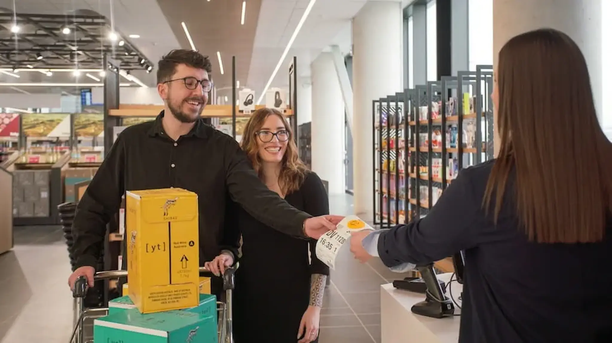 A couple at a store checkout counter, smiling as they hand a receipt to a cashier