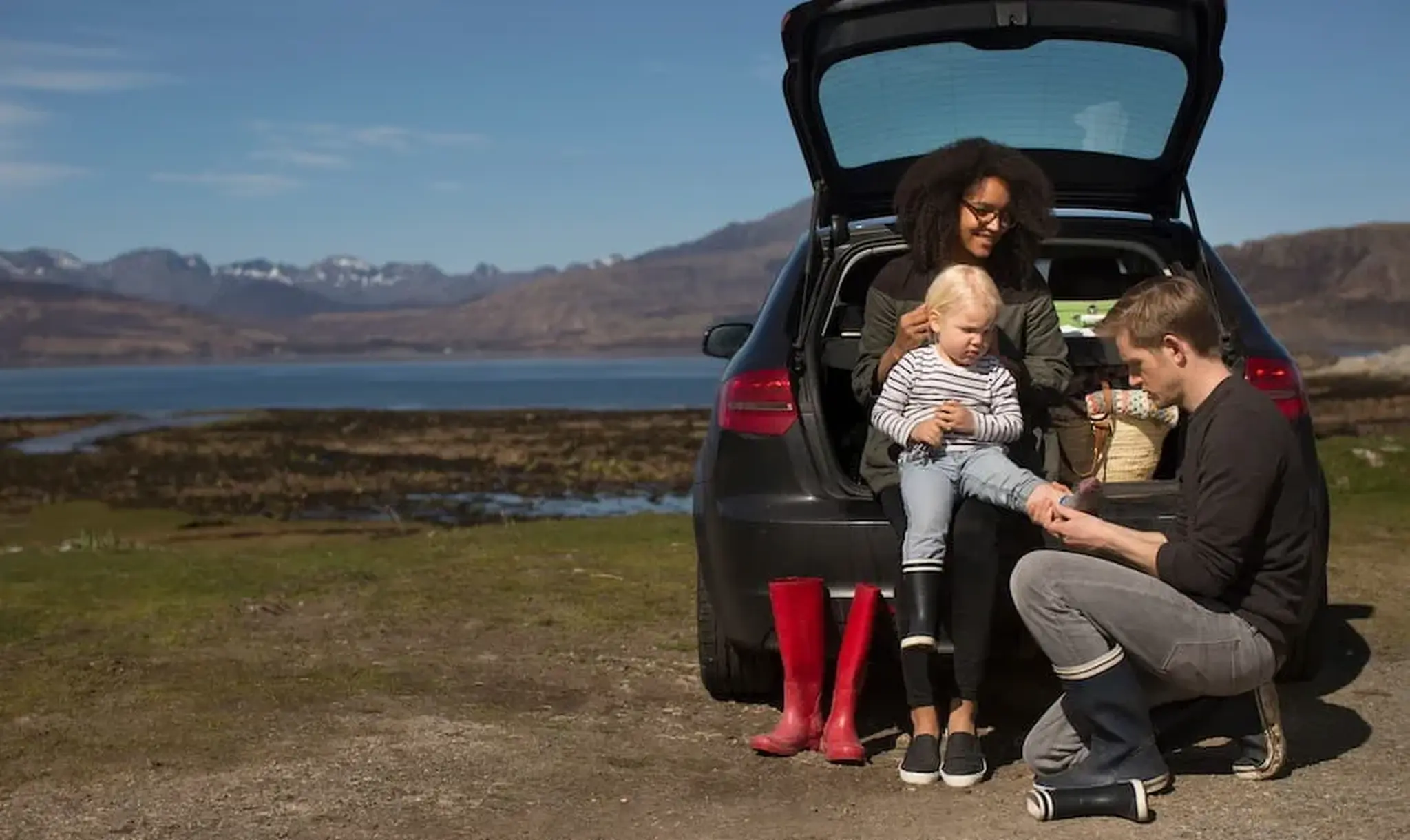 A family enjoys a scenic view by a lake, sitting at the open trunk of a car