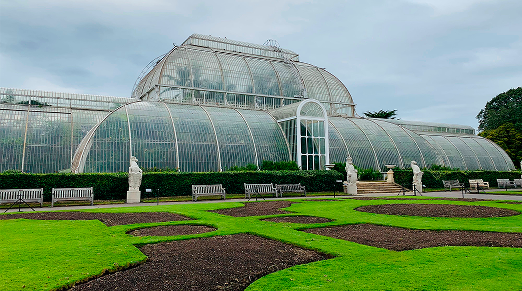 The Palm House at Kew Gardens with statues and green lawns under a cloudy sky.