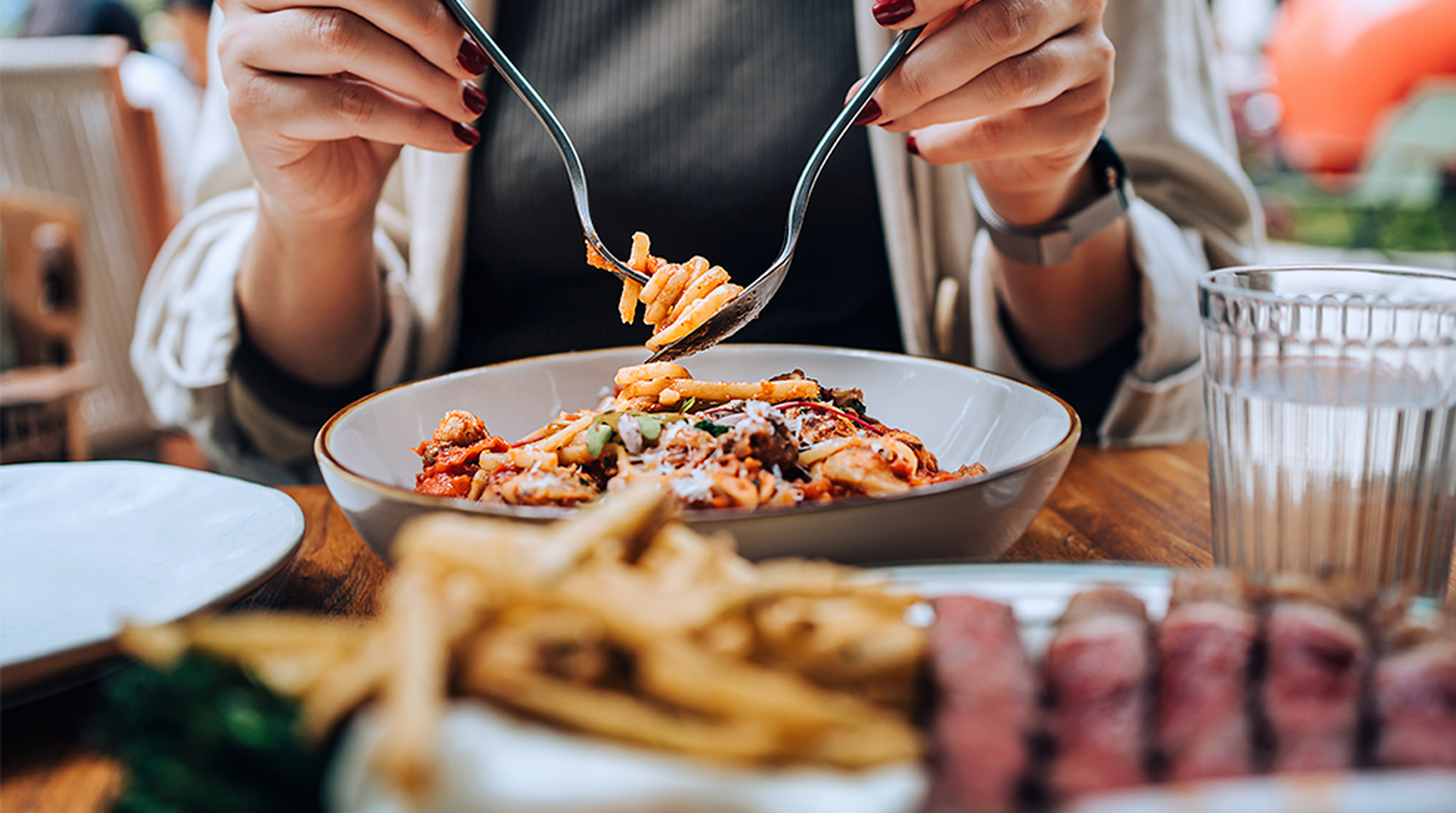 A person eating pasta with a fork, surrounded by a plate of fries, steak, and a glass of water