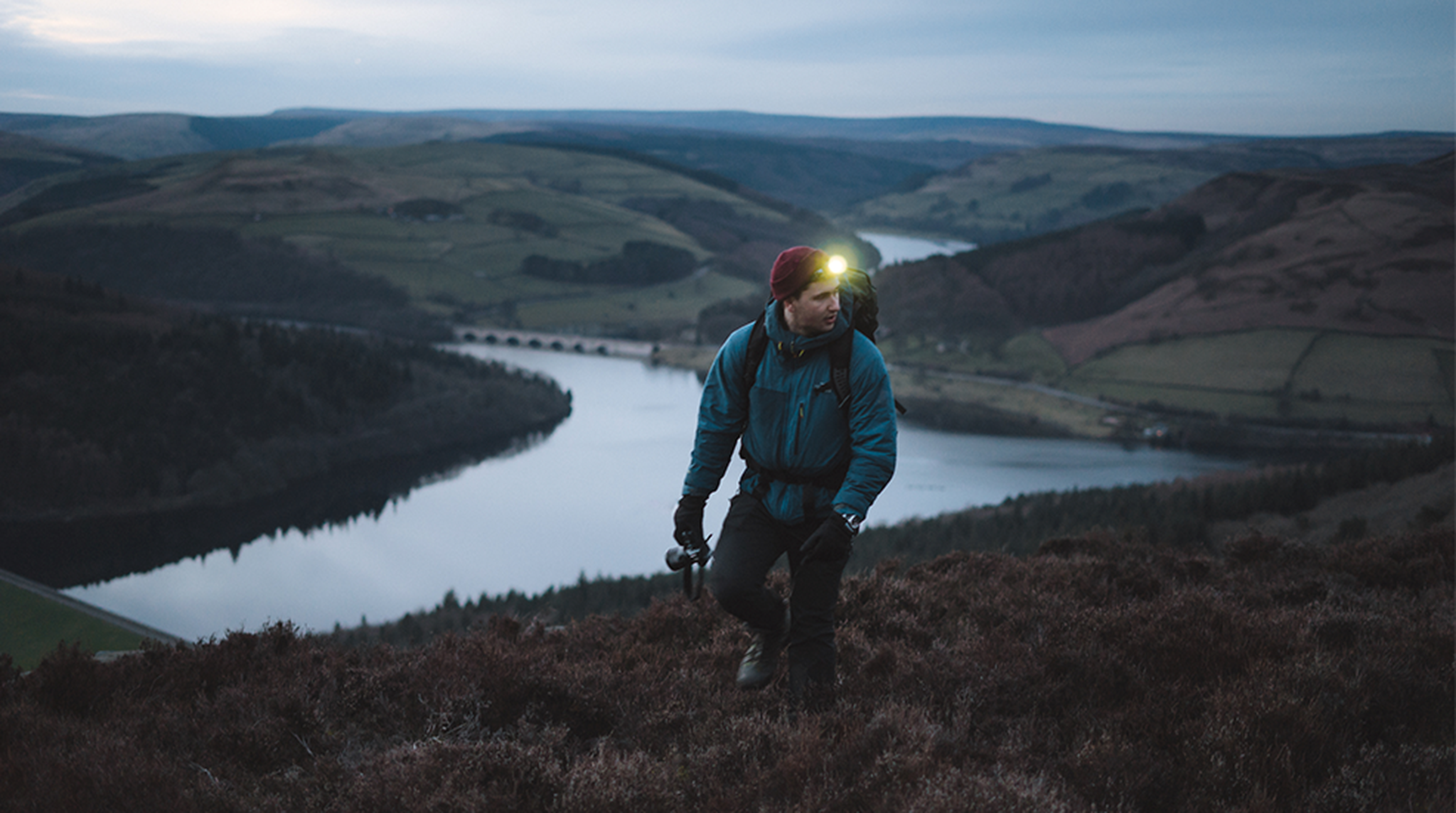 A hiker with a headlamp climbs a hill at dusk, overlooking a landscape of rolling hills and a river below.