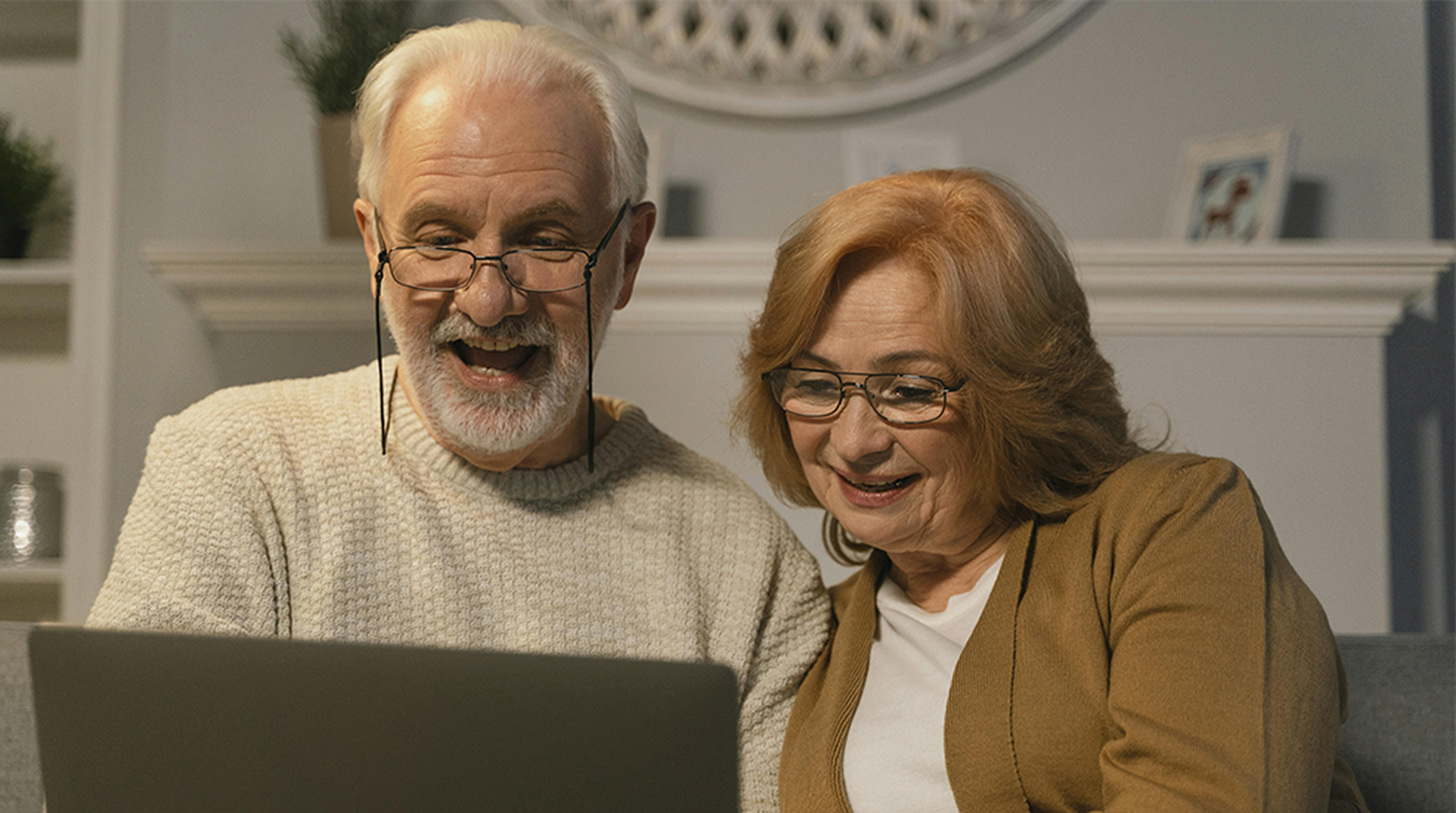 Elderly couple smiling while looking at a laptop on a sofa. Shelves with decor and a framed photo are in the background.