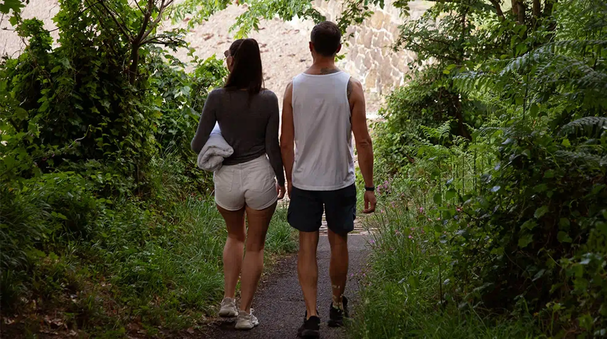 A couple walks along a narrow, tree-lined path, surrounded by greenery