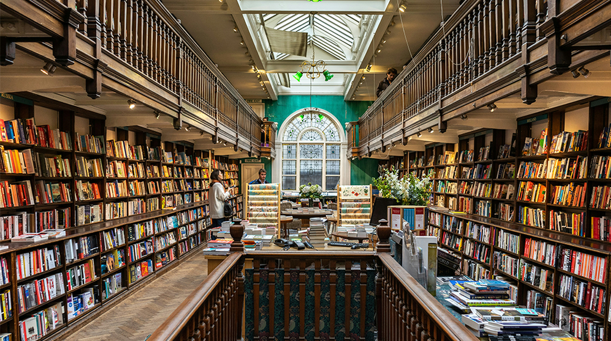 A cozy bookstore with tall shelves filled with books, a skylight above, and people browsing. A large arched window provides natural light.