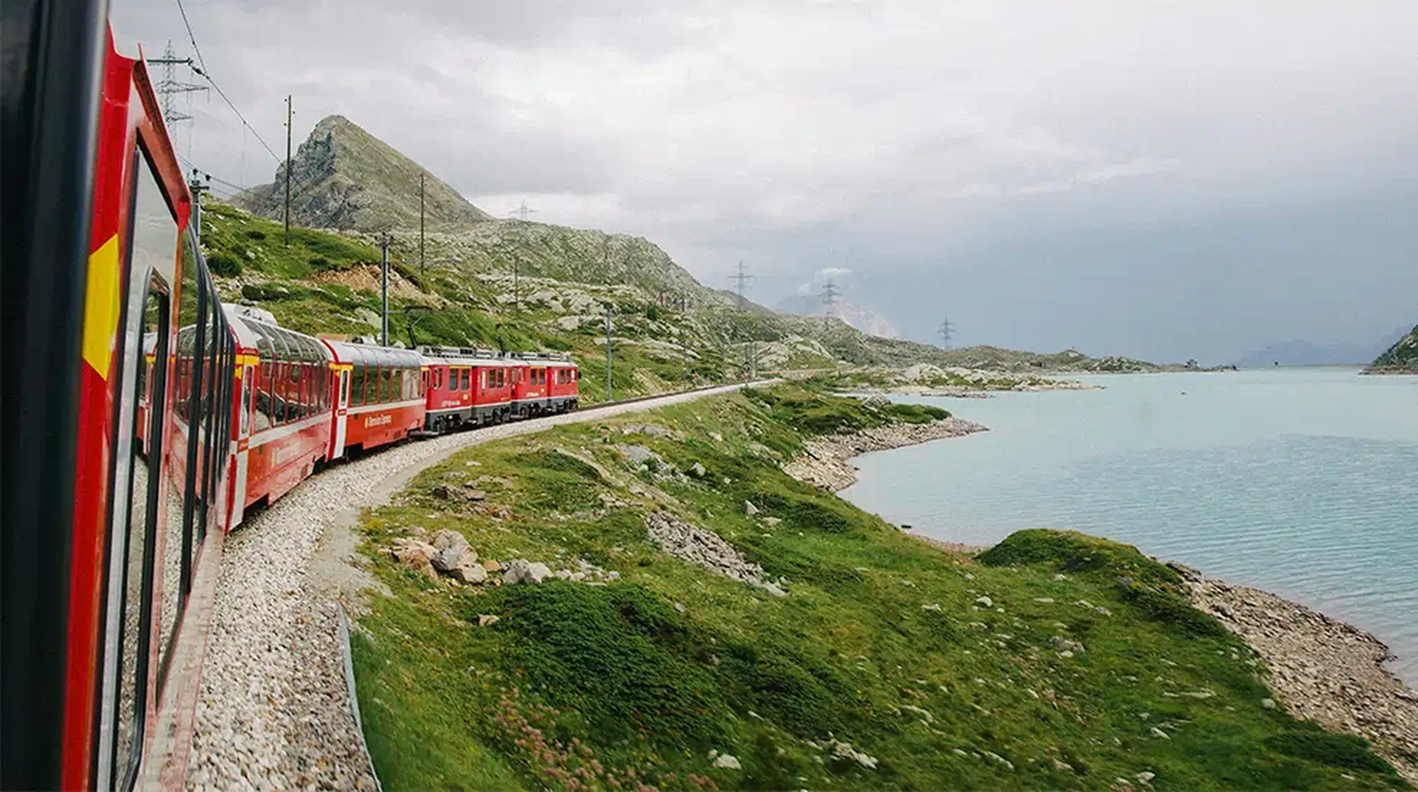 A red train travels along a curving track beside a turquoise lake and green mountains