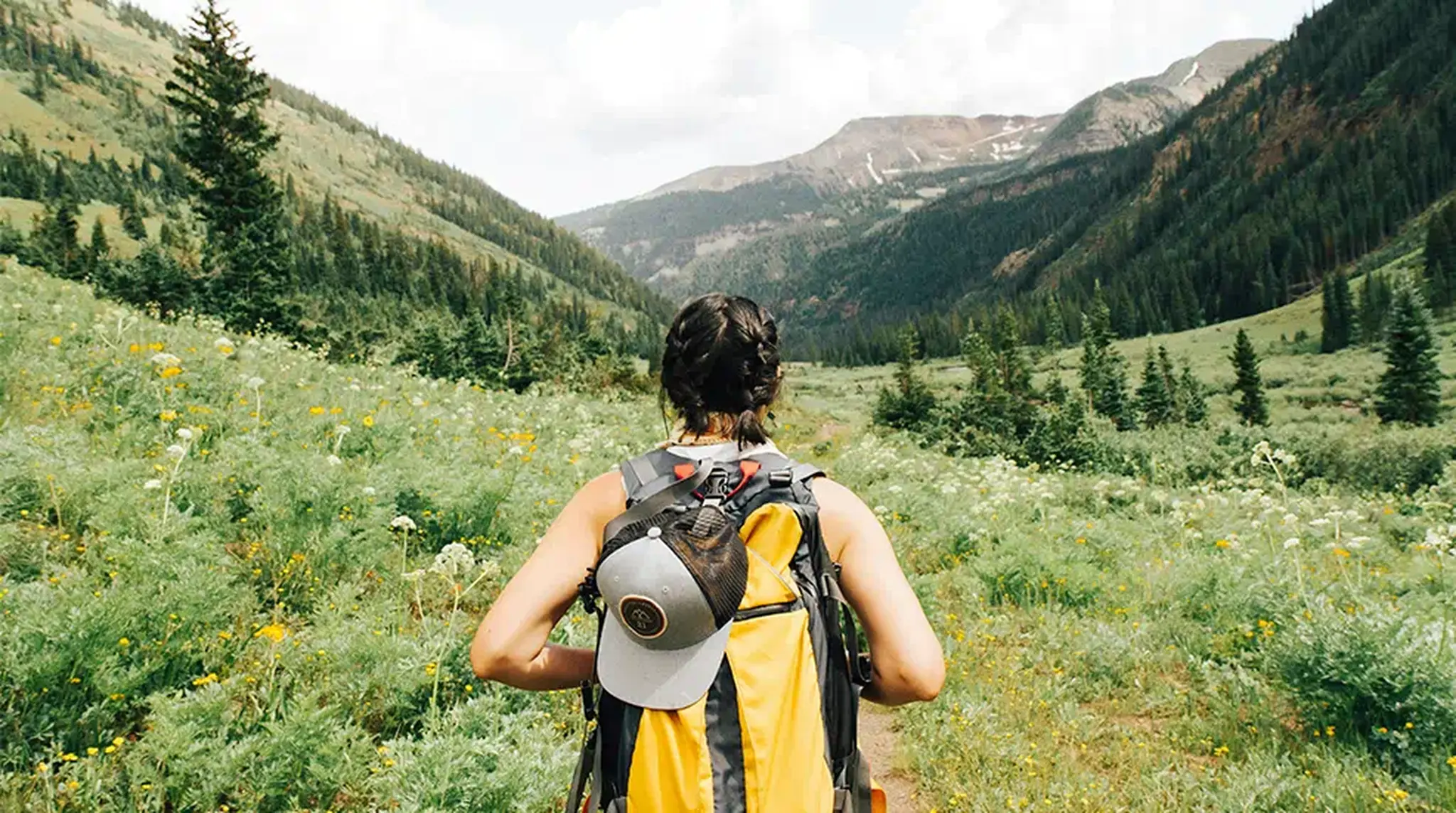 Hiker with a yellow backpack and cap walks through a mountain valley