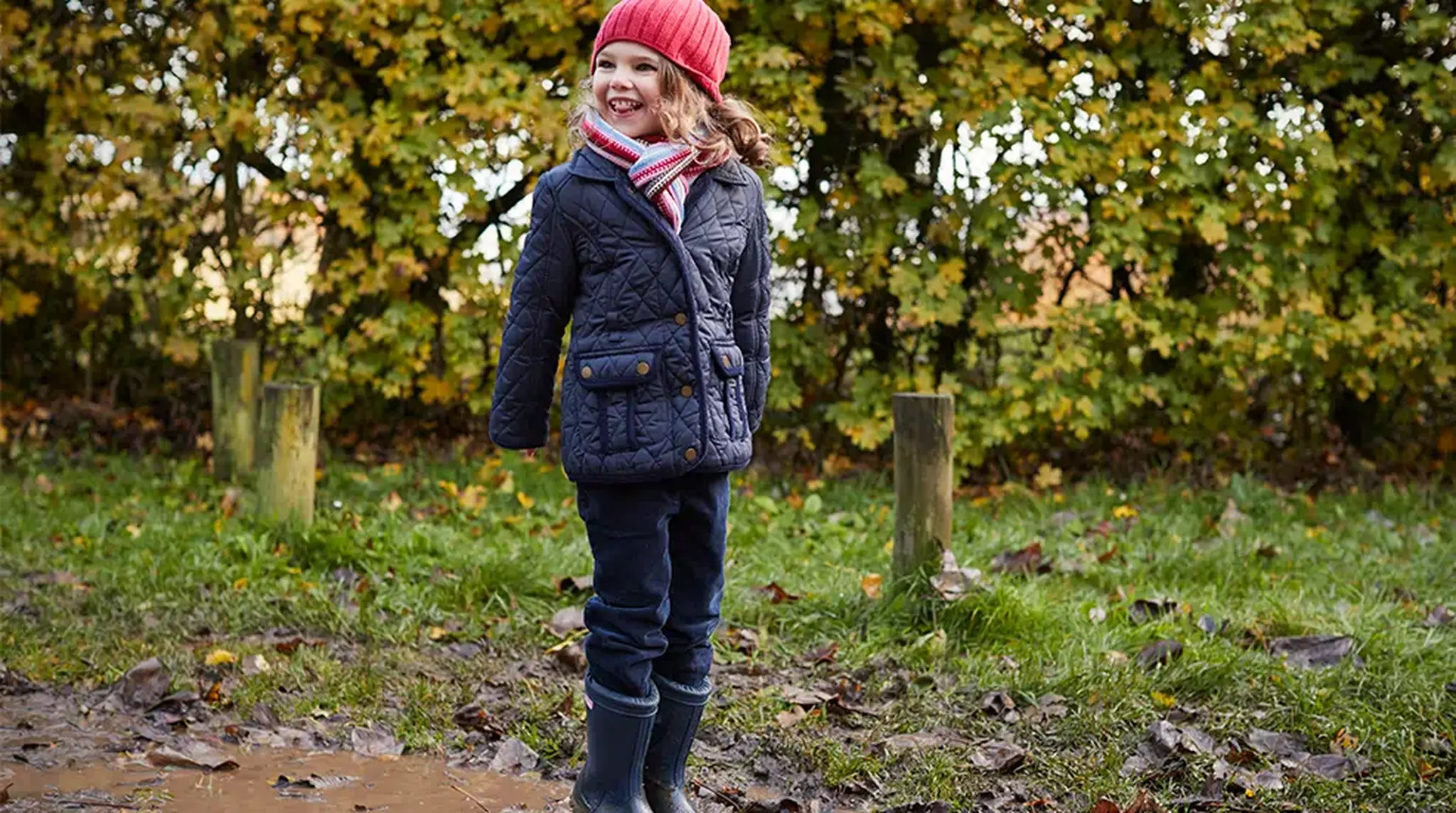 A young child in a red hat and scarf stands joyfully in a muddy puddle