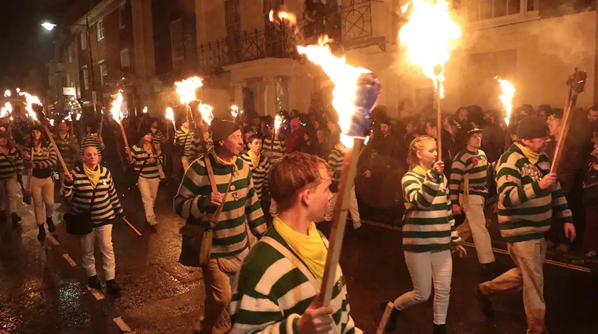People in striped shirts carry flaming torches during a night time parade on a street