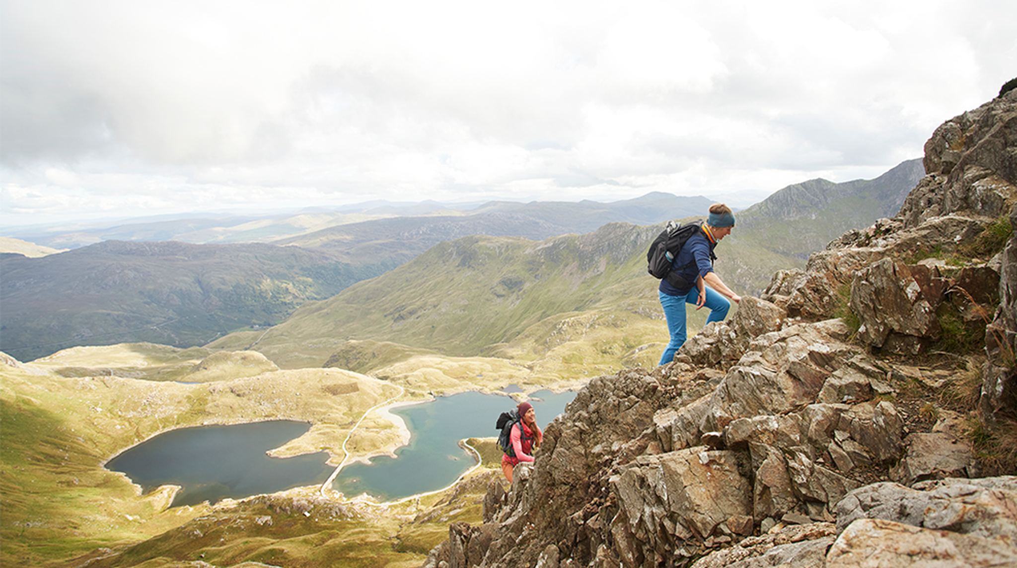 Hikers ascend a rocky mountain trail with panoramic views of lakes and rolling hills under a partly cloudy sky.