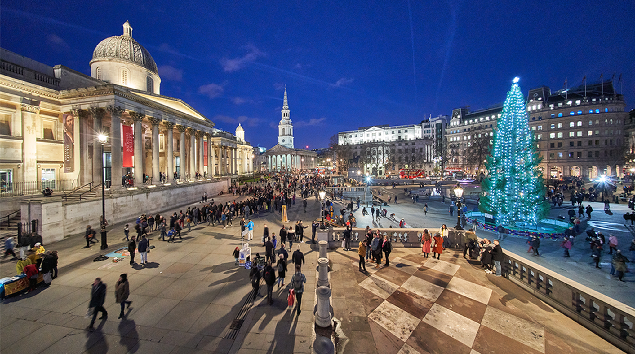 A bustling evening at Trafalgar Square with a tall, illuminated Christmas tree, lit buildings, and crowds of people enjoying the scene.