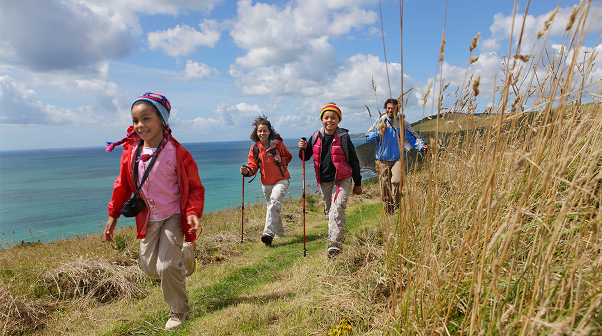 A family of four hikes on a grassy coastal trail with trekking poles, overlooking the ocean under a partly cloudy sky.