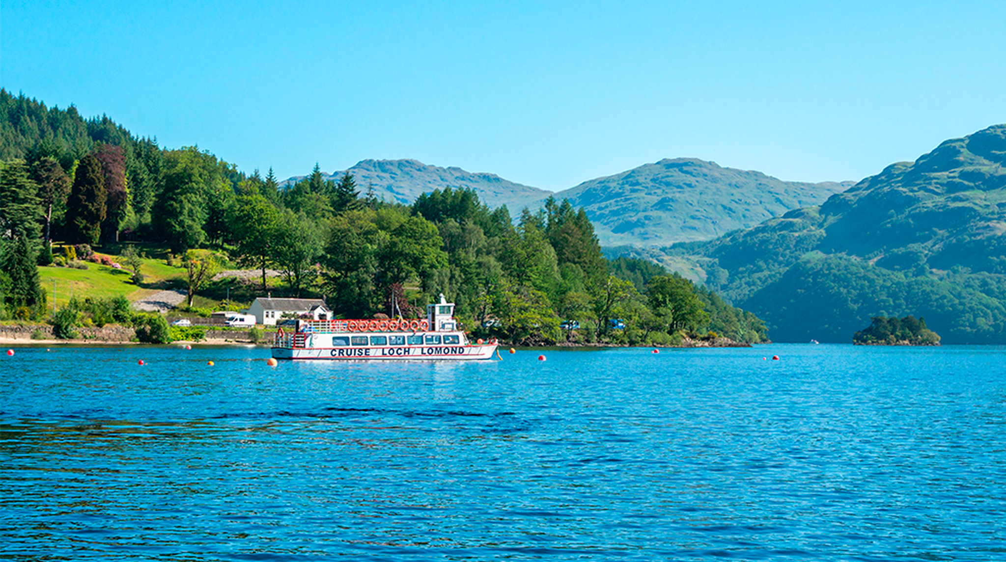 Tourists pack a boat that is cruising on Loch Lomond, on a bright, clear blue sky day, with forest and green tree filled mountains in the background