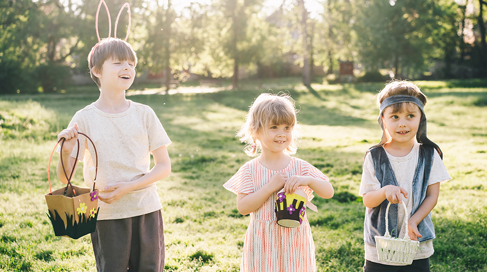 Three children in a sunny park, two holding baskets and one wearing bunny ears, smile during an Easter egg hunt.