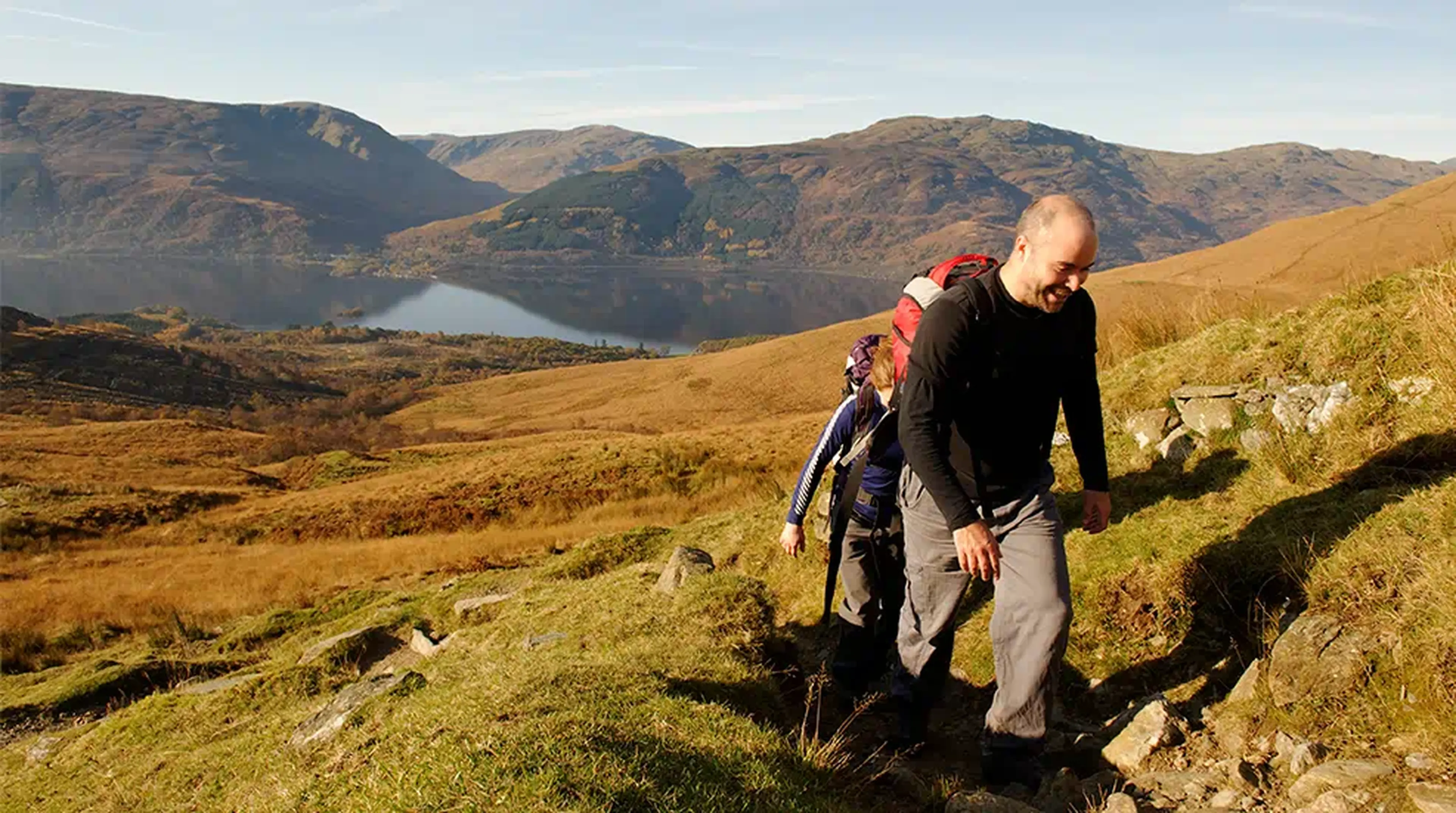 Two hikers ascend a grassy trail with backpacks, overlooking a serene lake and mountainous landscape