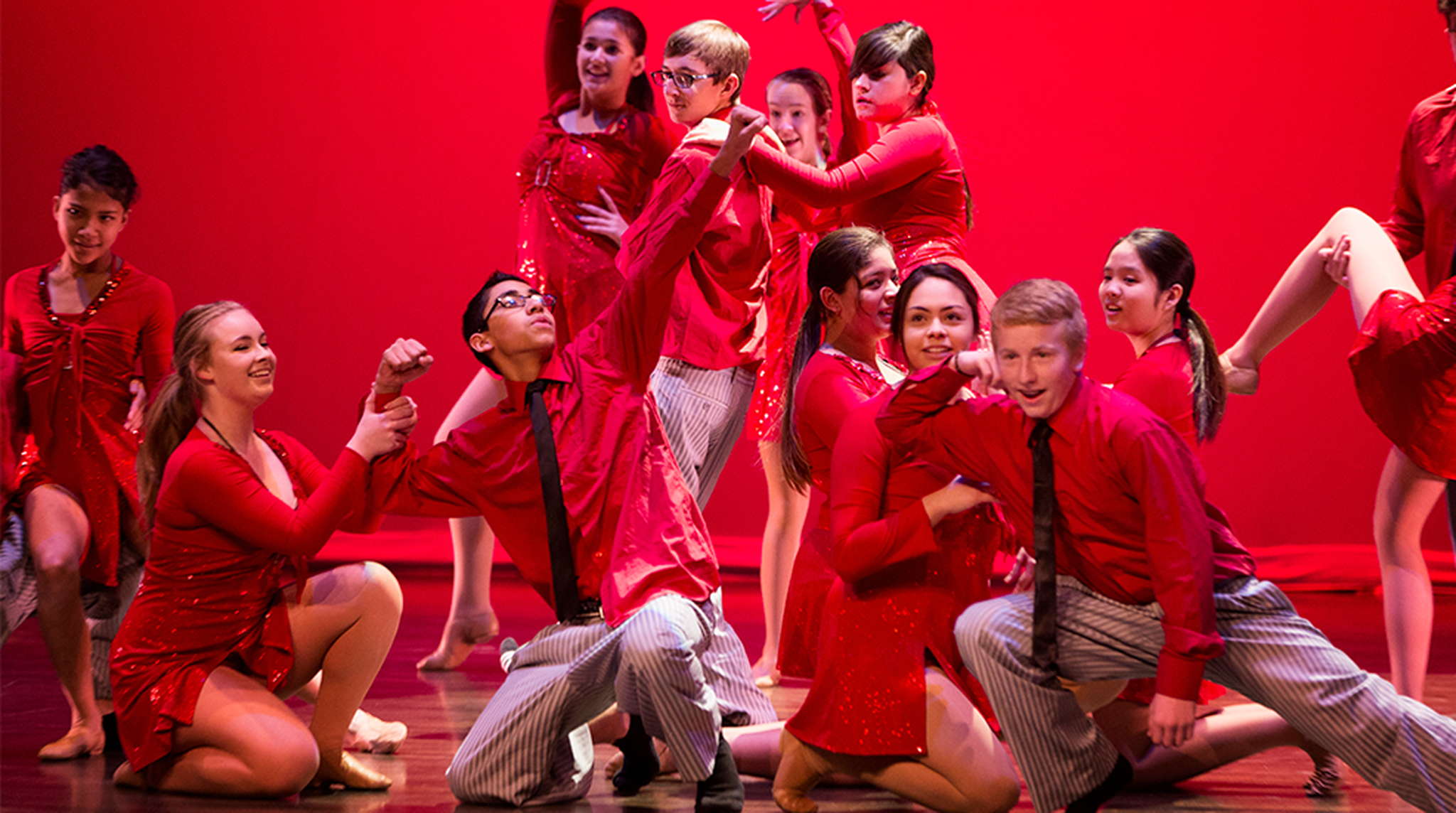 A group of performers in red costumes pose energetically on stage with a vibrant red background