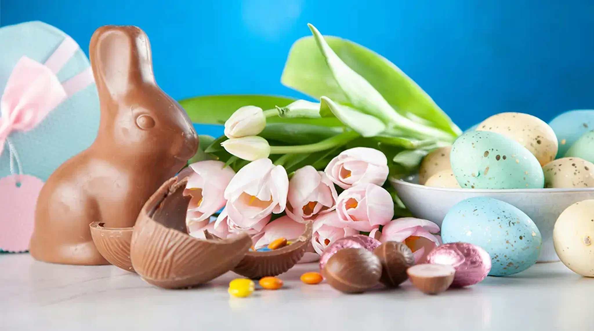 A chocolate bunny, broken chocolate egg, pastel-colored Easter eggs, pink tulips
