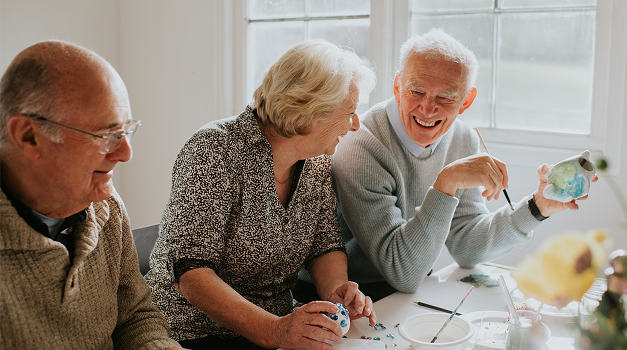 Three older adults sit around a table by a sunlit window, sharing a cheerful moment while working on small ceramic crafts
