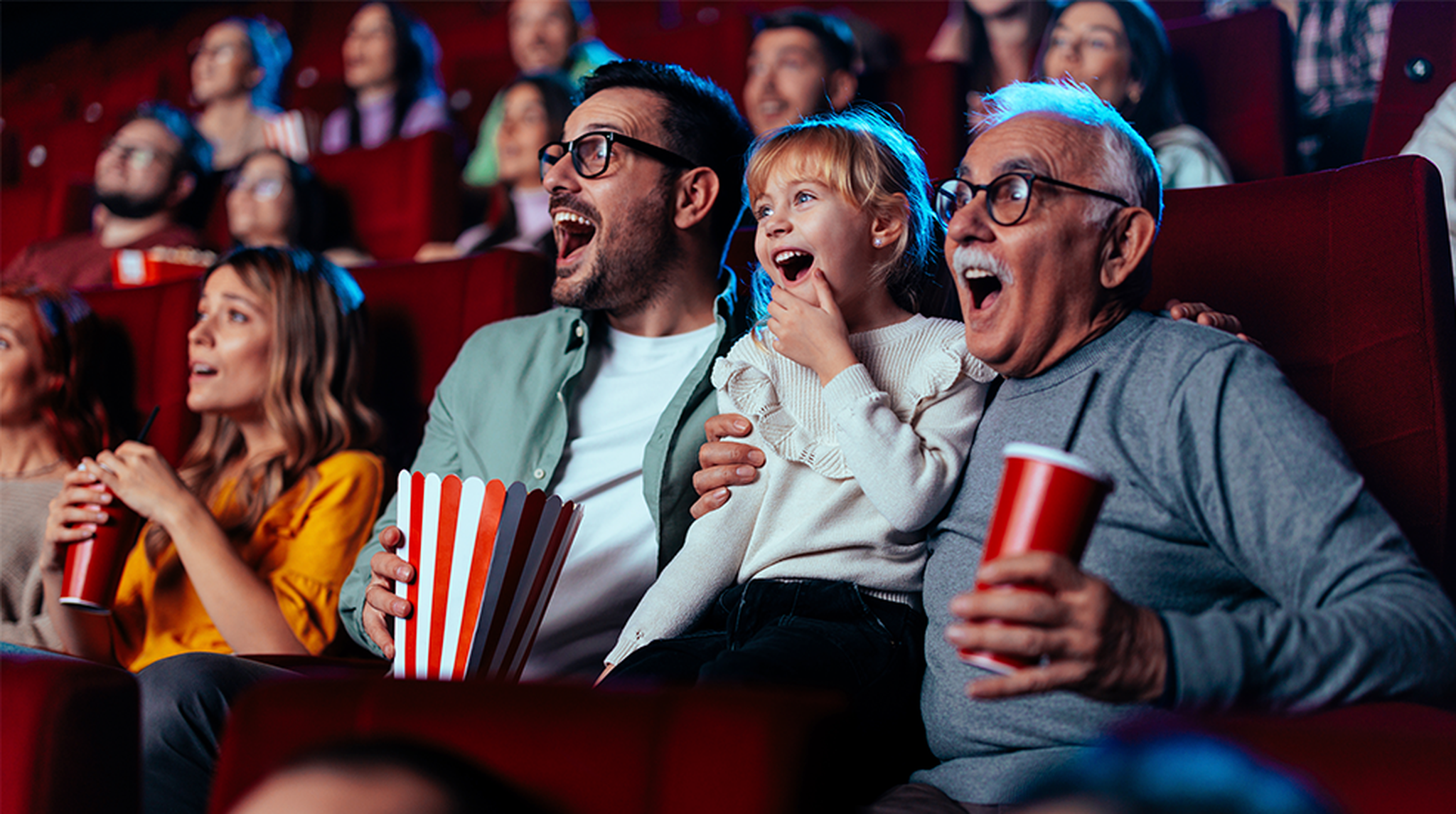 Three people, including a child, sit in a theater, looking surprised. They hold popcorn and drinks, surrounded by other engaged audience members.