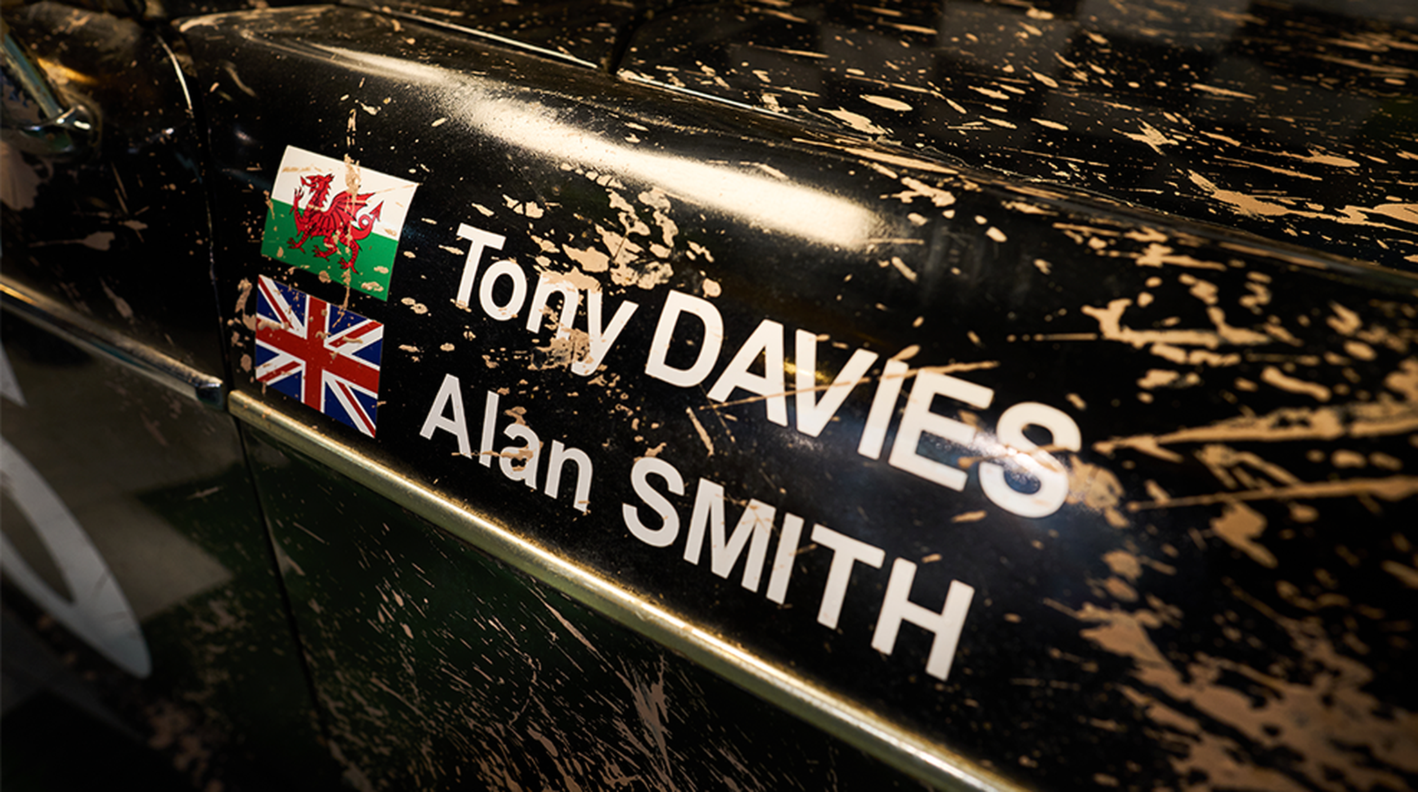 Close-up of a car's side, showing Welsh and British flags with the names Tony Davies and Alan Smith amidst mud splatters.