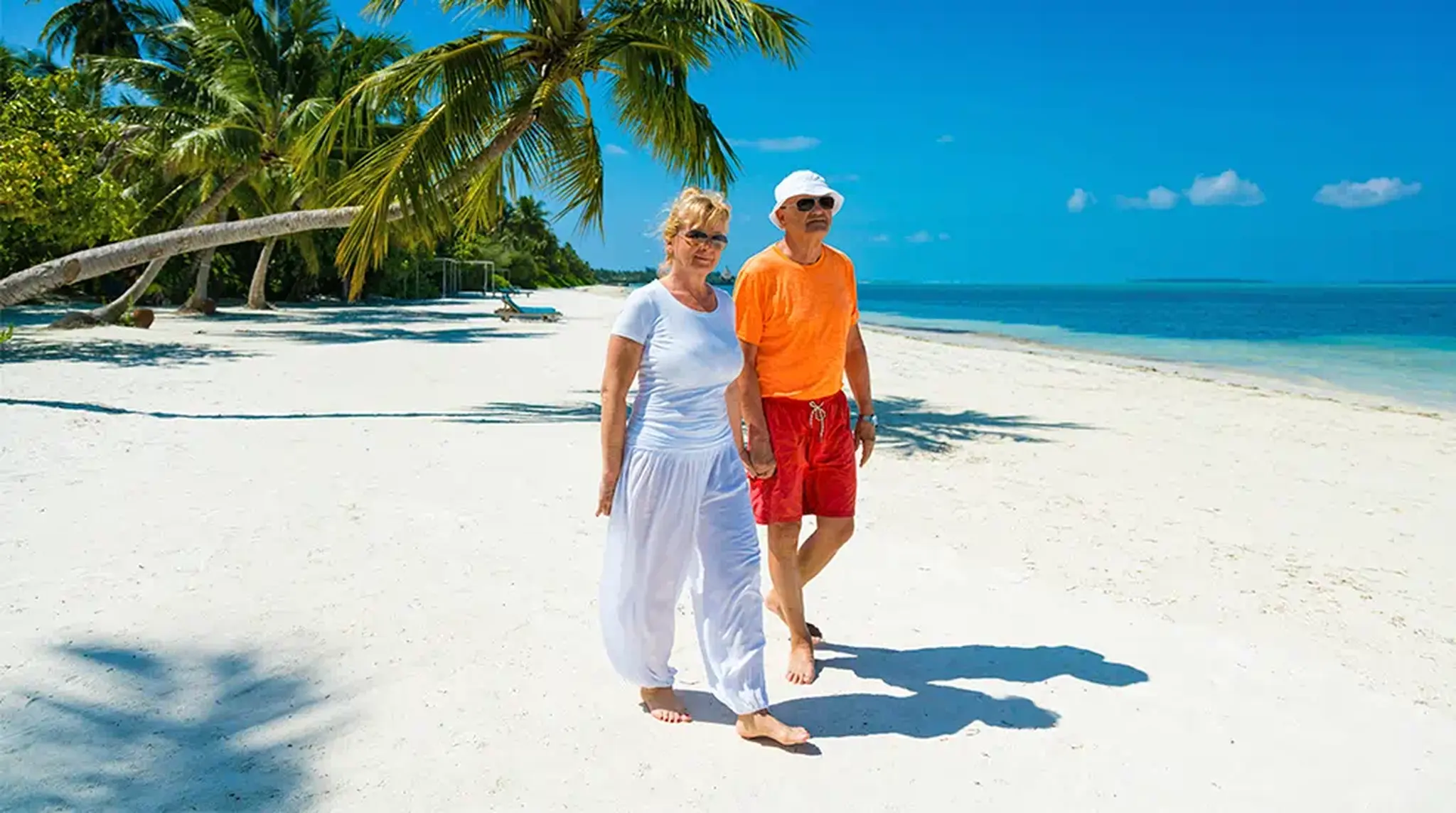An elderly couple walks barefoot on a sunny beach beside clear blue water