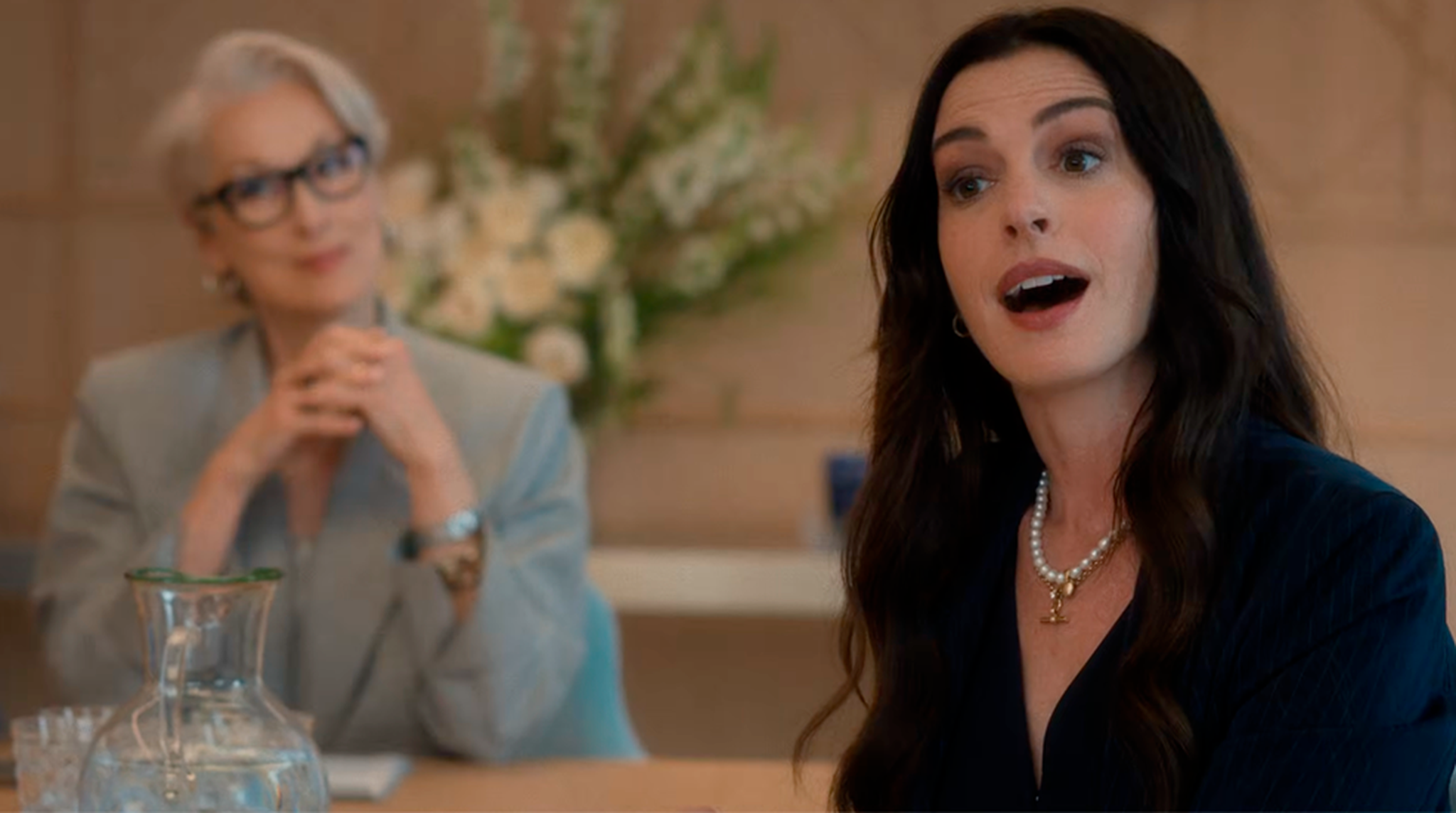 Two women are in a professional setting. One is speaking enthusiastically, while the other listens attentively. Flowers and a water jug adorn the table.
