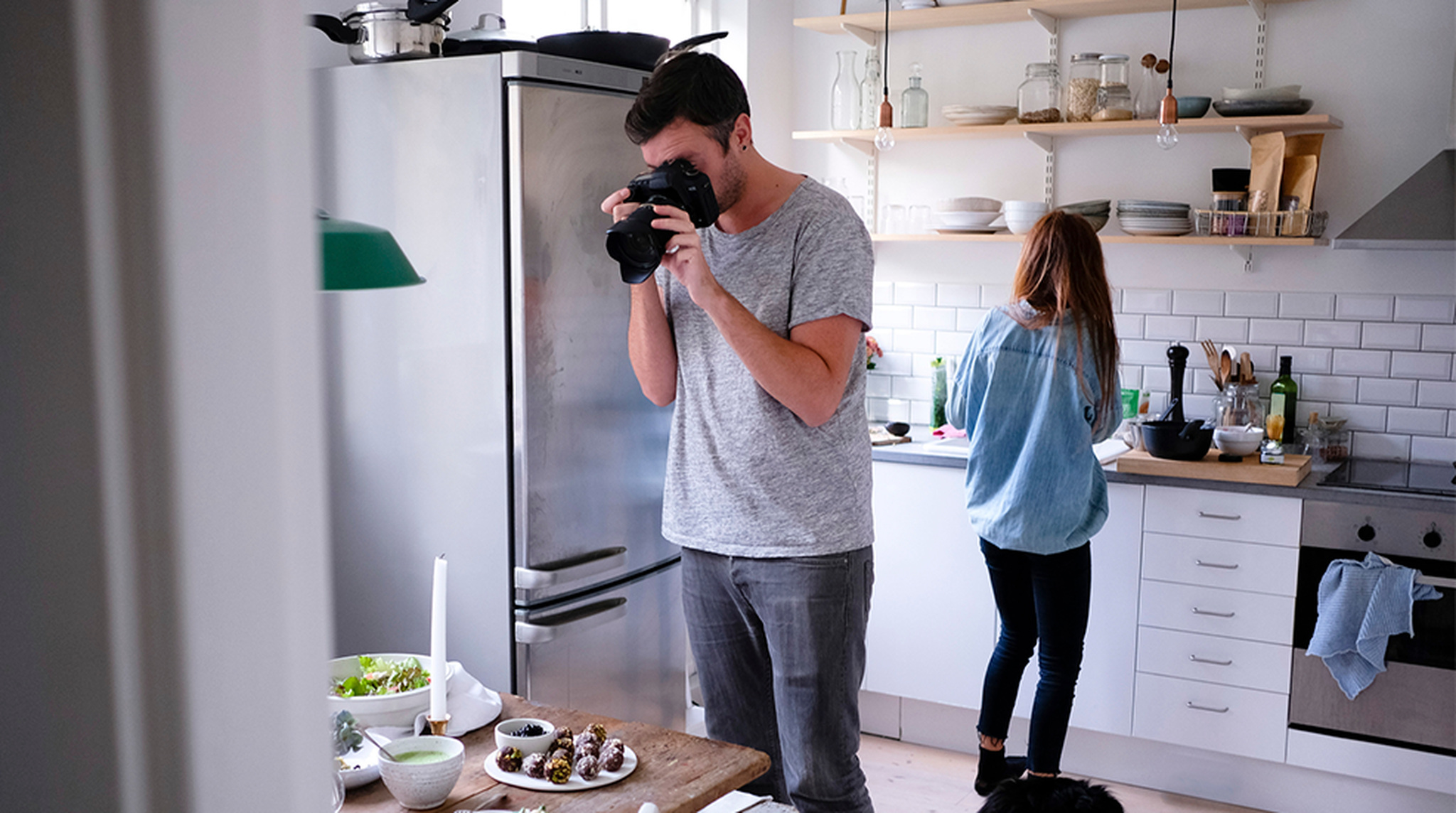 A couple in the kitchen, the man holding a large camera to his face and taking a photo of the food they're preparing