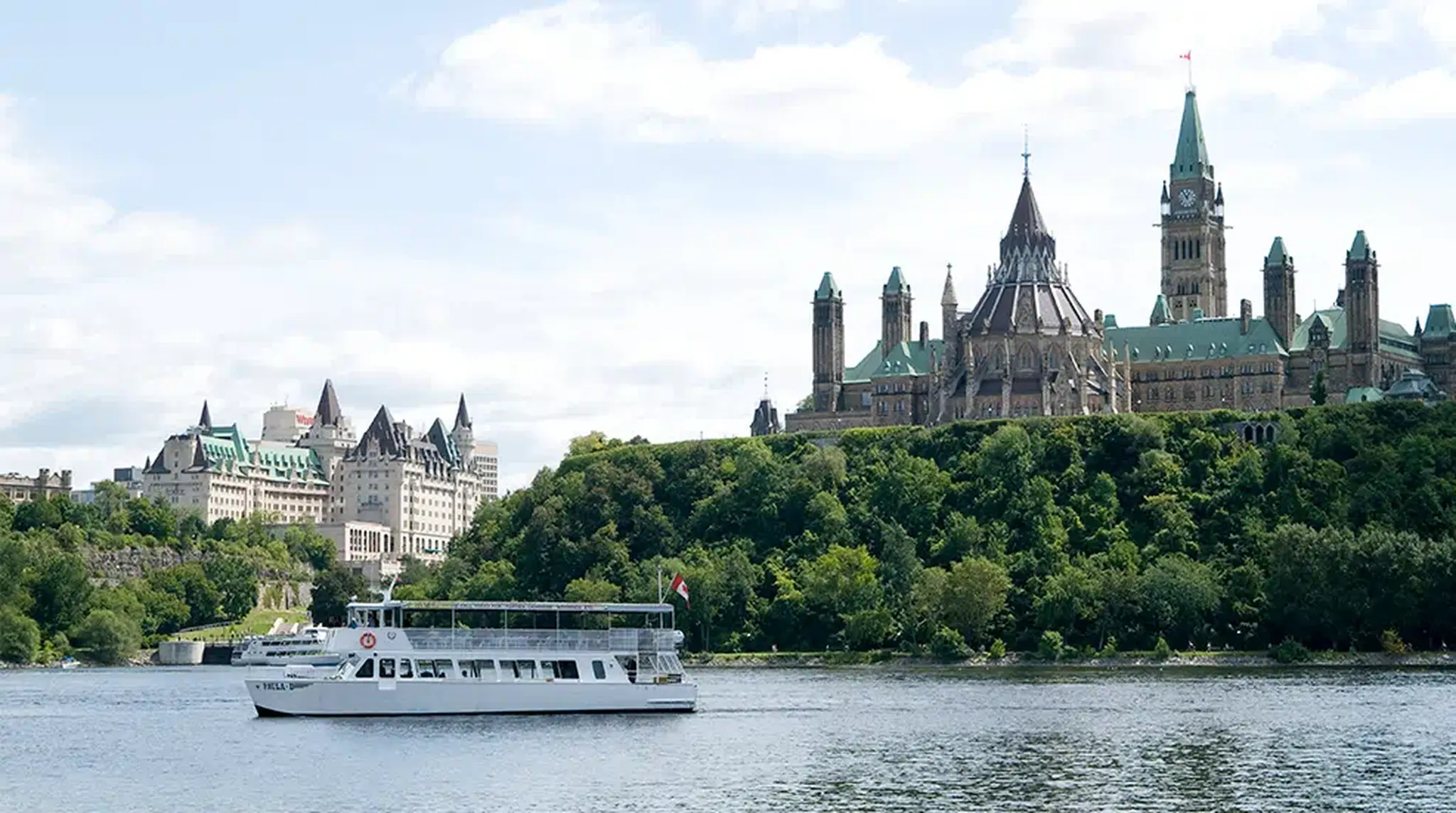 A tour boat sails on a river with the Gothic-style buildings of Ottawa's Parliament Hill in the background