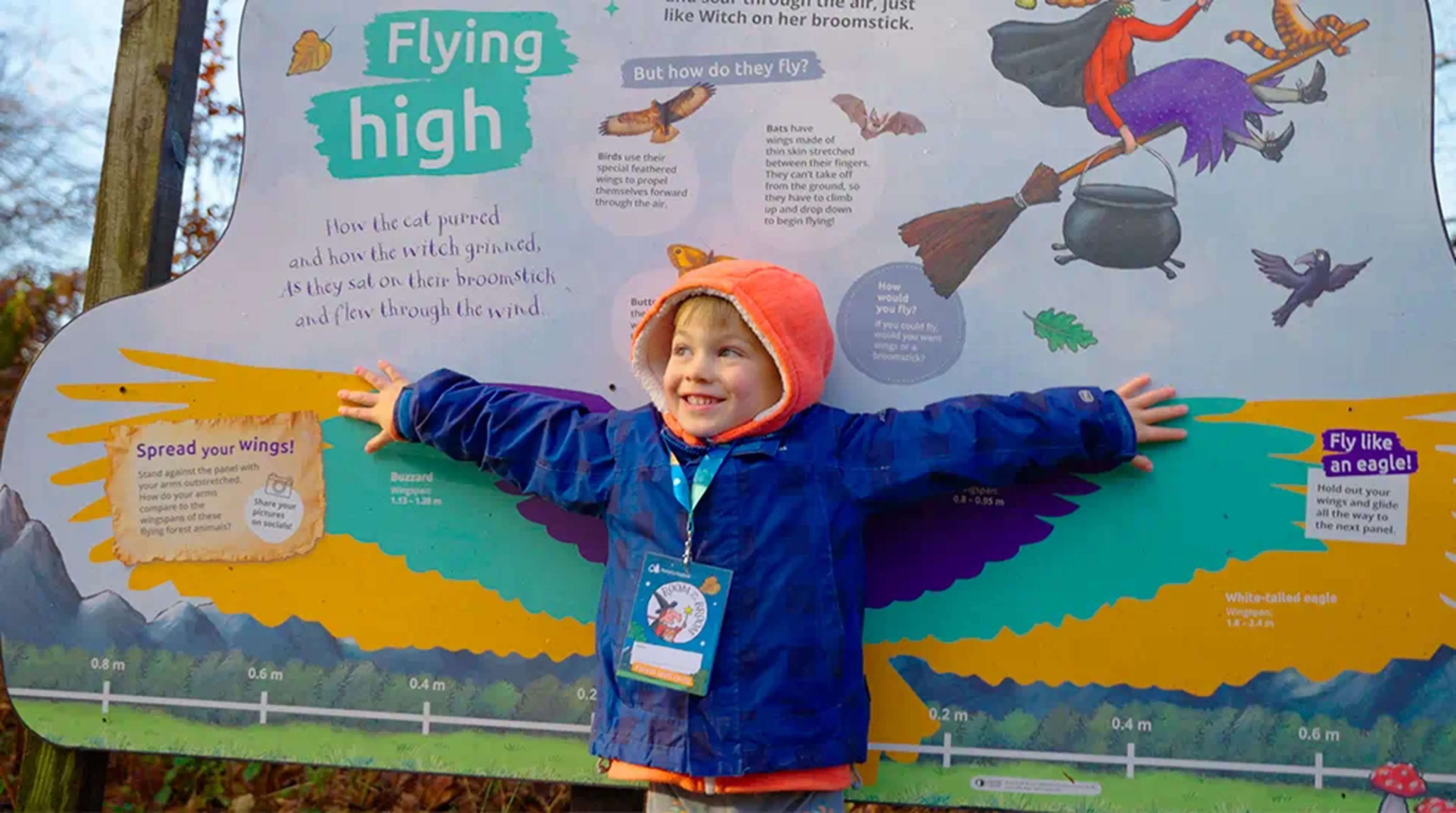 A smiling child in an orange hood stands with arms outstretched in front of a colourful educational sign about flying