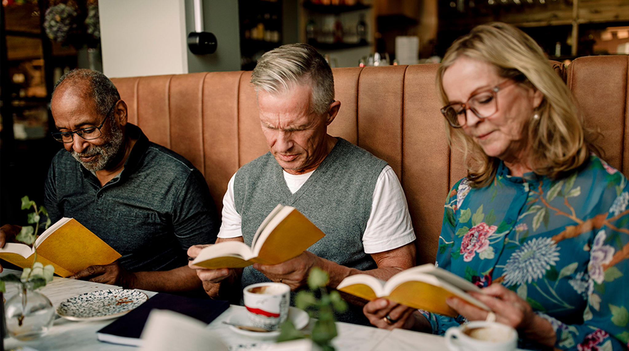 Three adults sitting in a café booth, reading books with yellow covers. They have beverages on the table and appear focused.