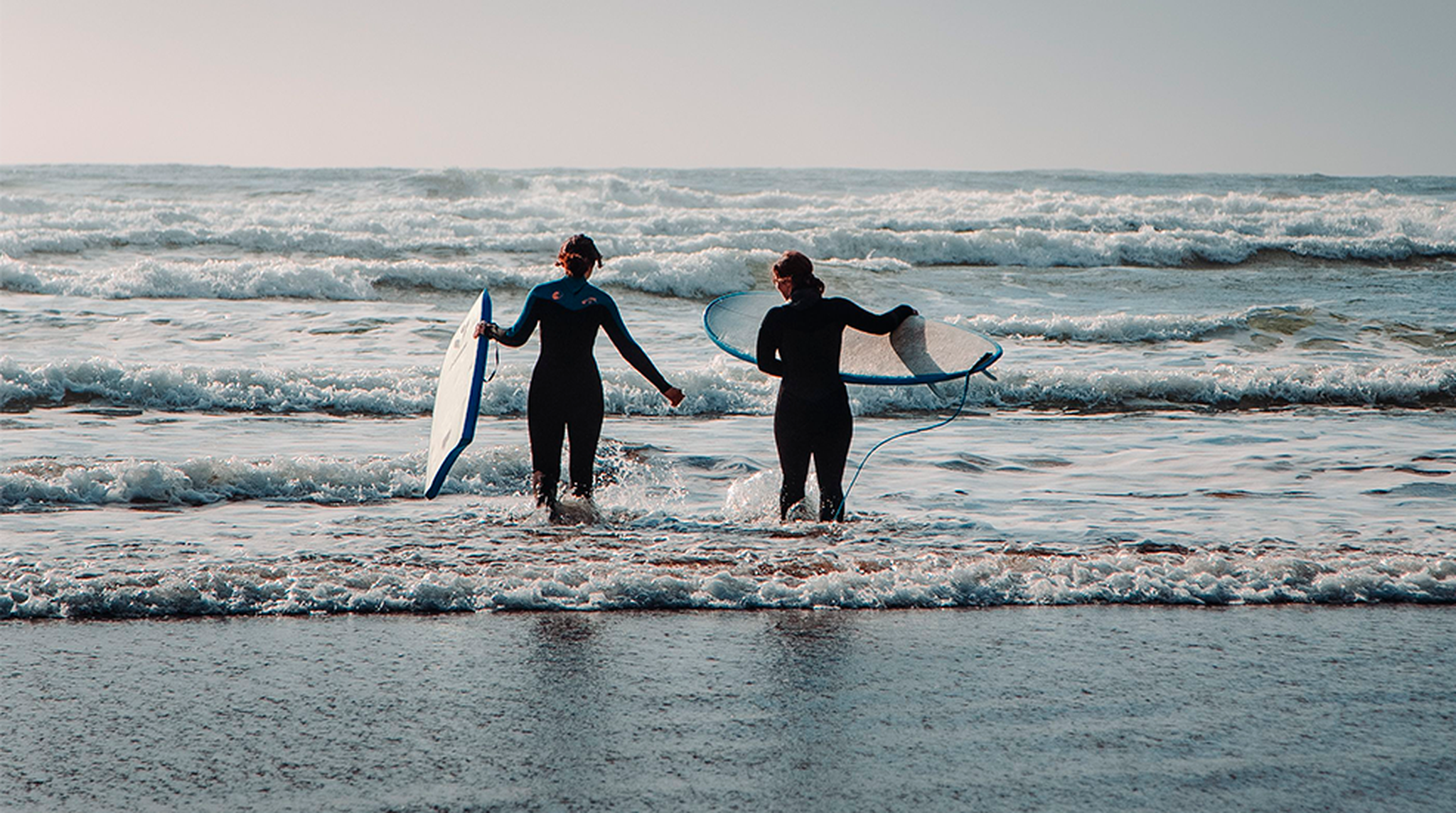 Two surfers with surfboards head into an overcast ocean with gentle waves.