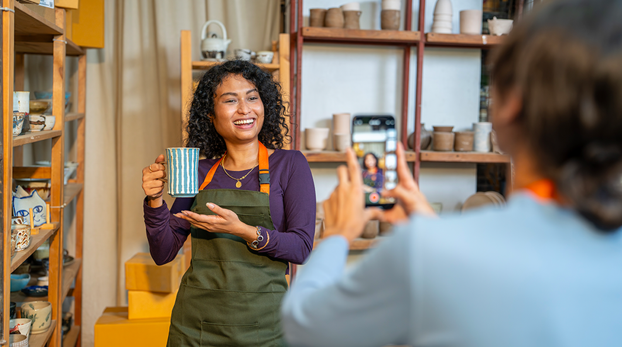 Smiling female ceramist holds mug while being filmed in workshop