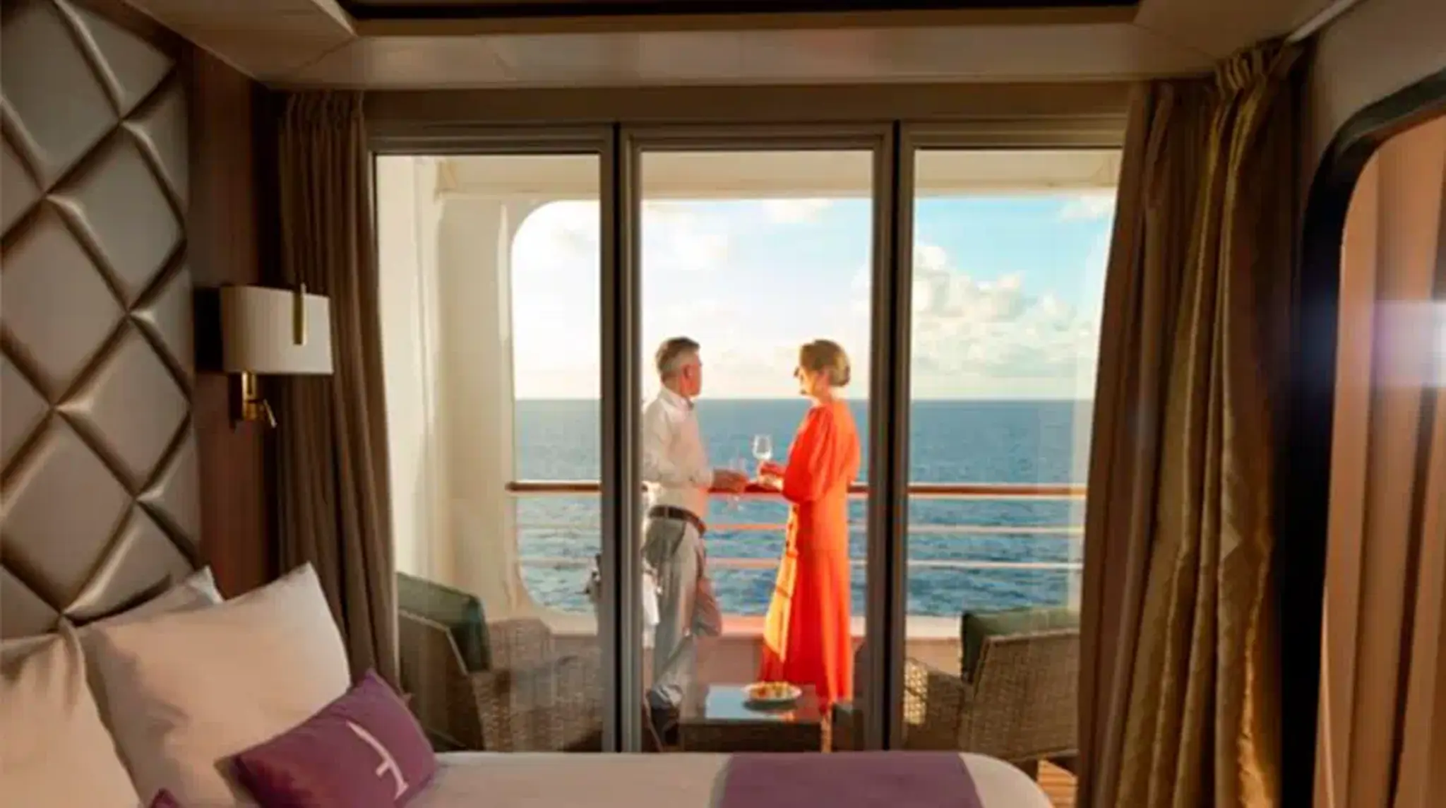 A couple stands on a cruise ship balcony, holding drinks and looking at the ocean