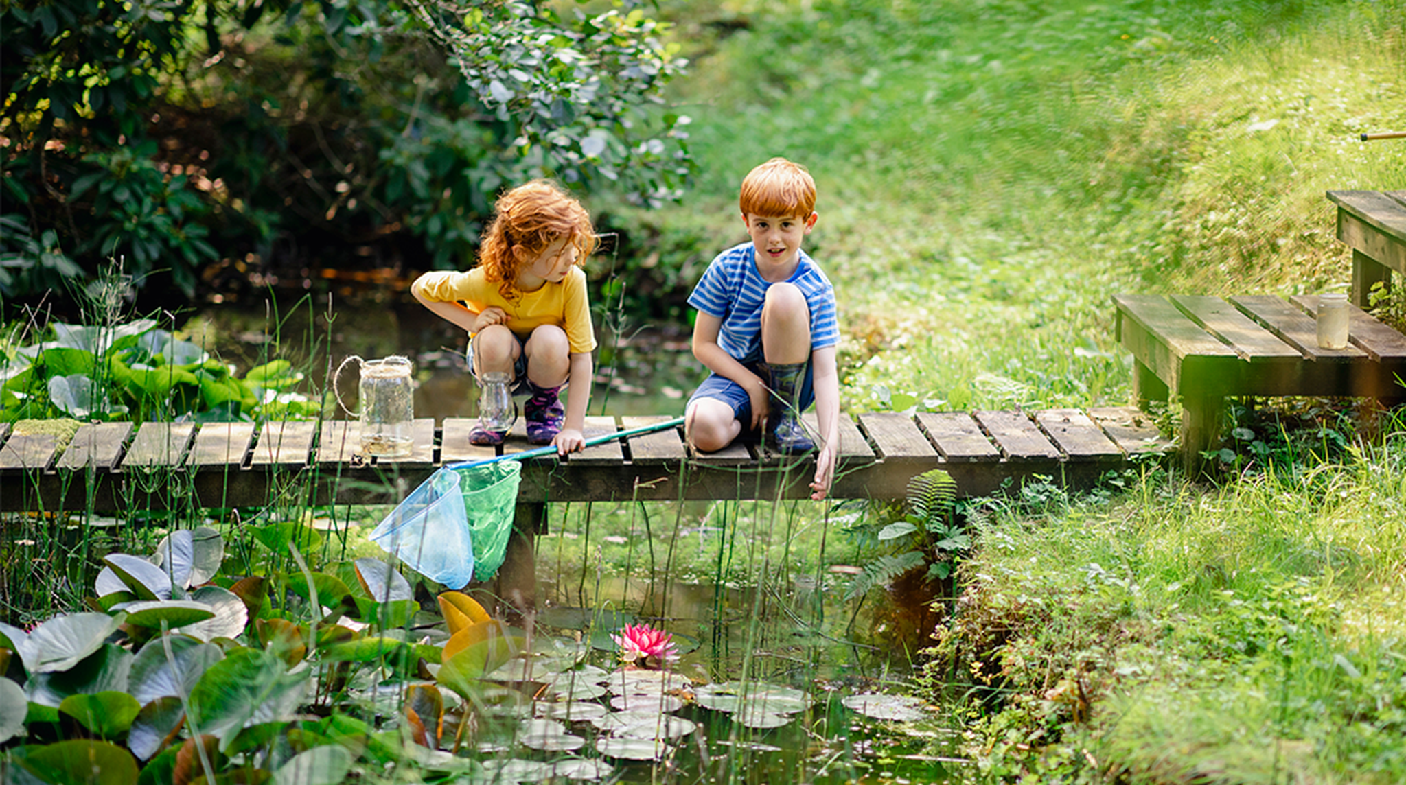 Young redhead brother and sister holding butterfly fishing nets crouching on a wooden platform over a pond, ready to go dipping.