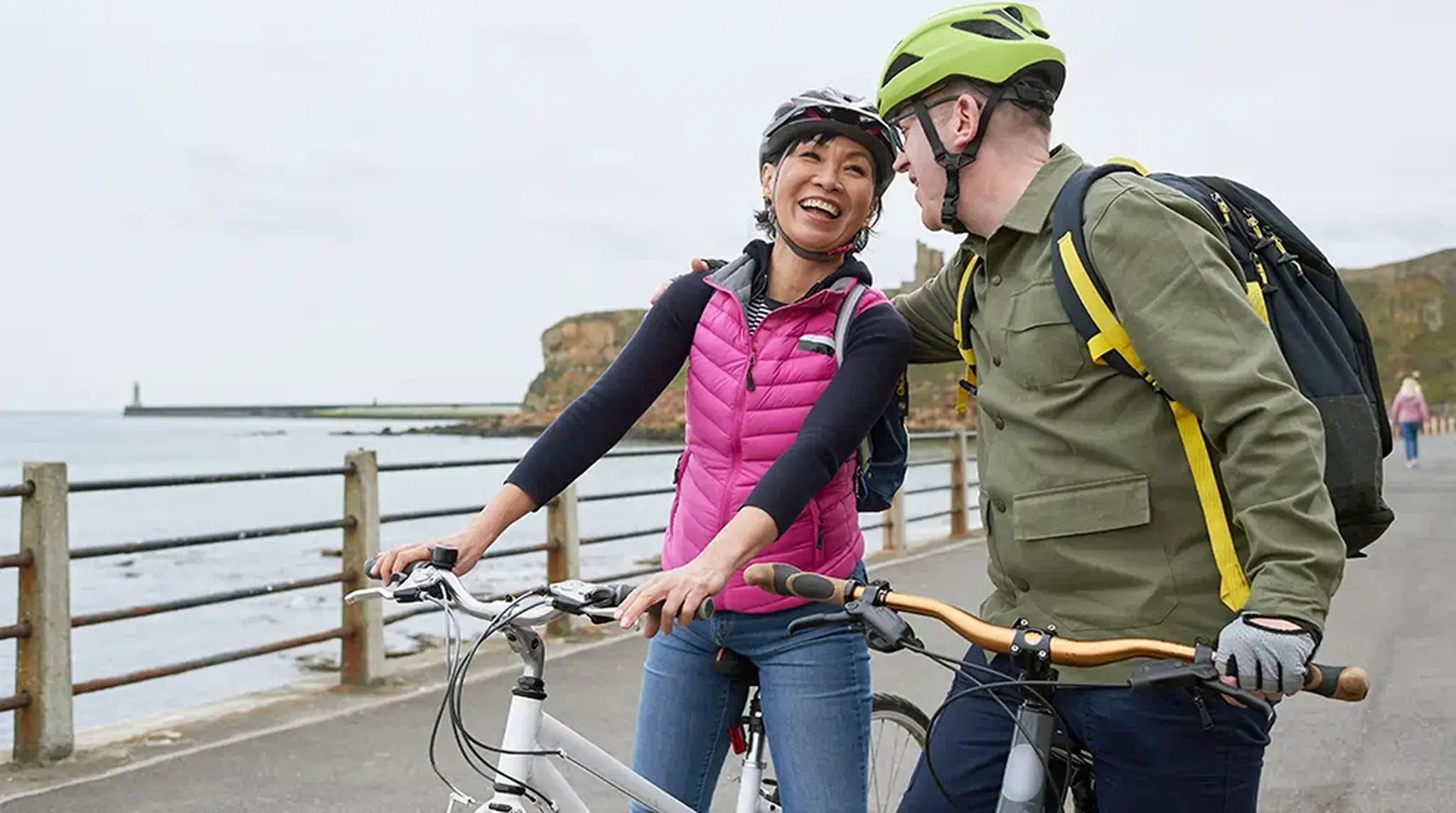 A woman and man in helmets smile while standing with their bikes on a seaside path