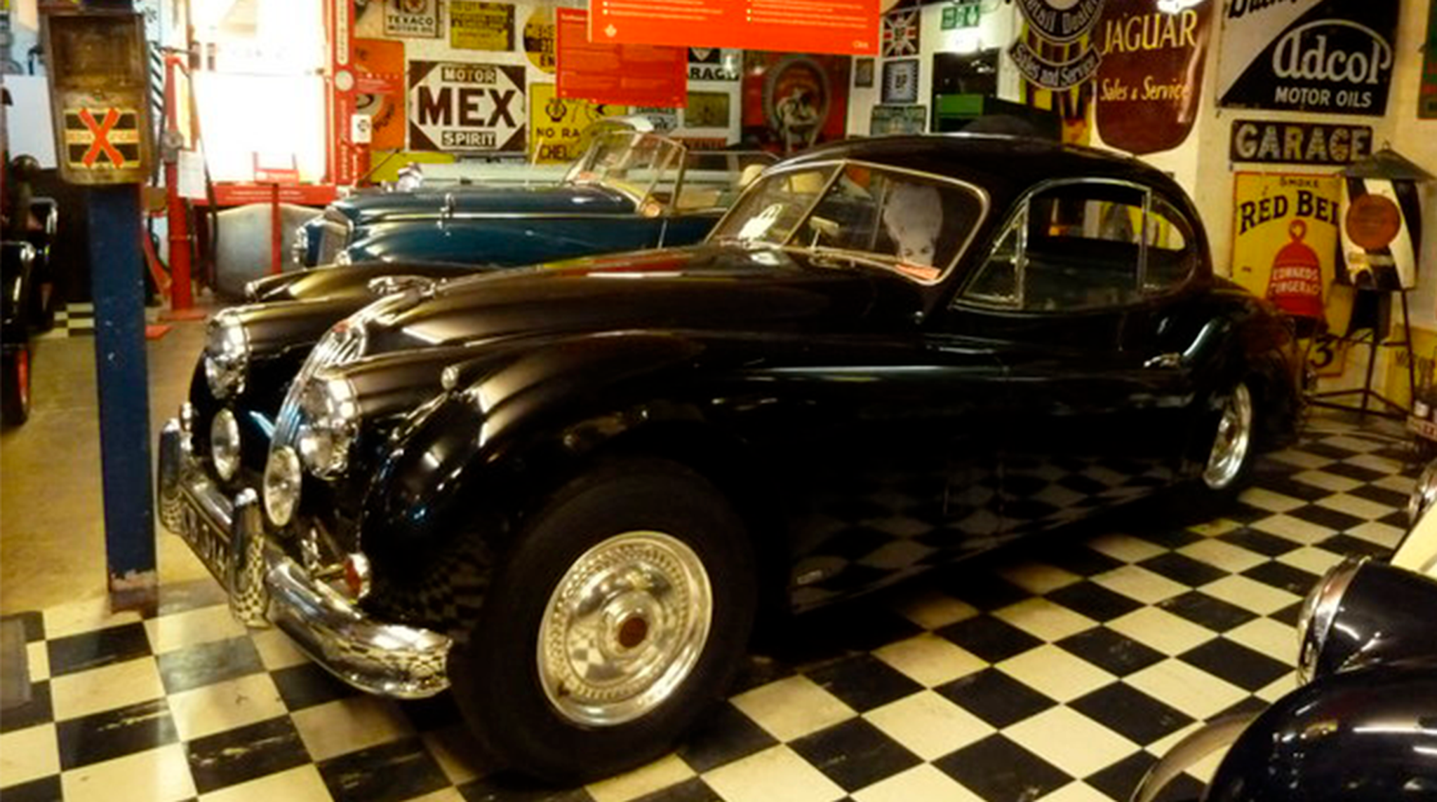 A vintage black car with a glossy finish displayed in a retro-themed garage with checkered flooring and vintage signs.