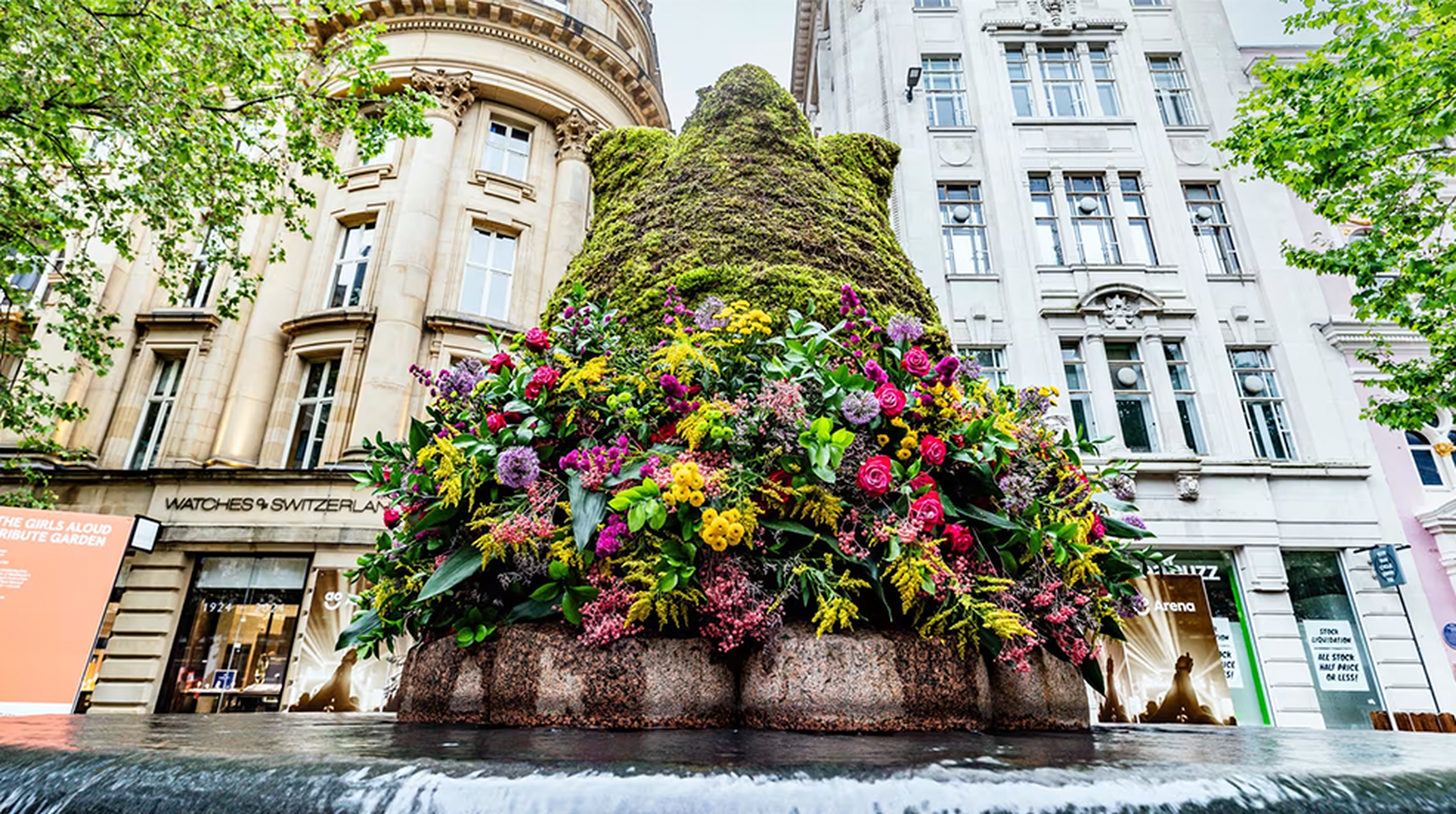 Bust of a moss-covered bear adorned with vibrant flowers in front of ornate buildings and a city street scene.