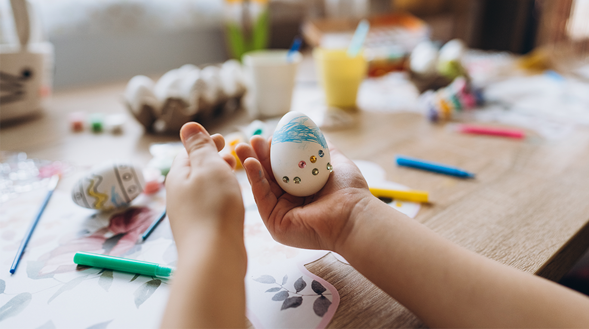 Child decorating an Easter egg with blue marker and rhinestones on a wooden table, surrounded by craft supplies and other decorated eggs.