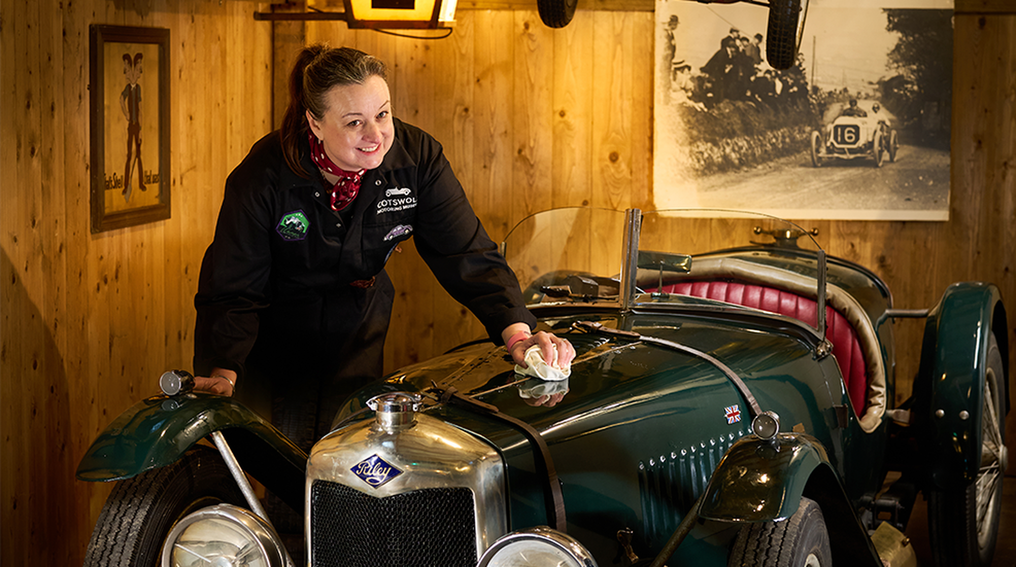 Woman in a black jacket polishes a vintage green car in a wooden garage with racing memorabilia on the walls.